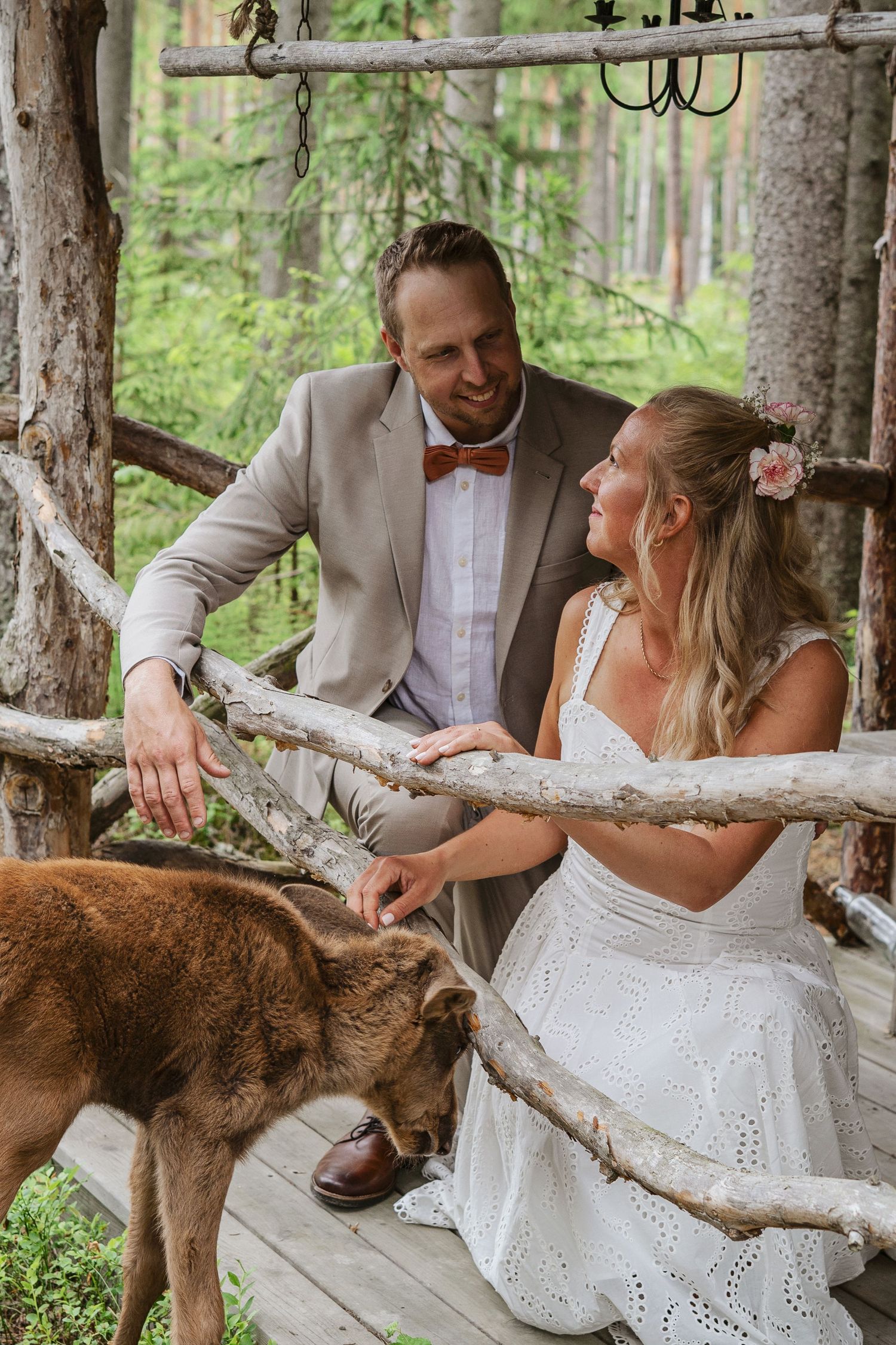 Brudpar poserar tillsammans med älgkalven vid en rustik träbyggnad under sin bröllopsfotografering i Ockelbo.