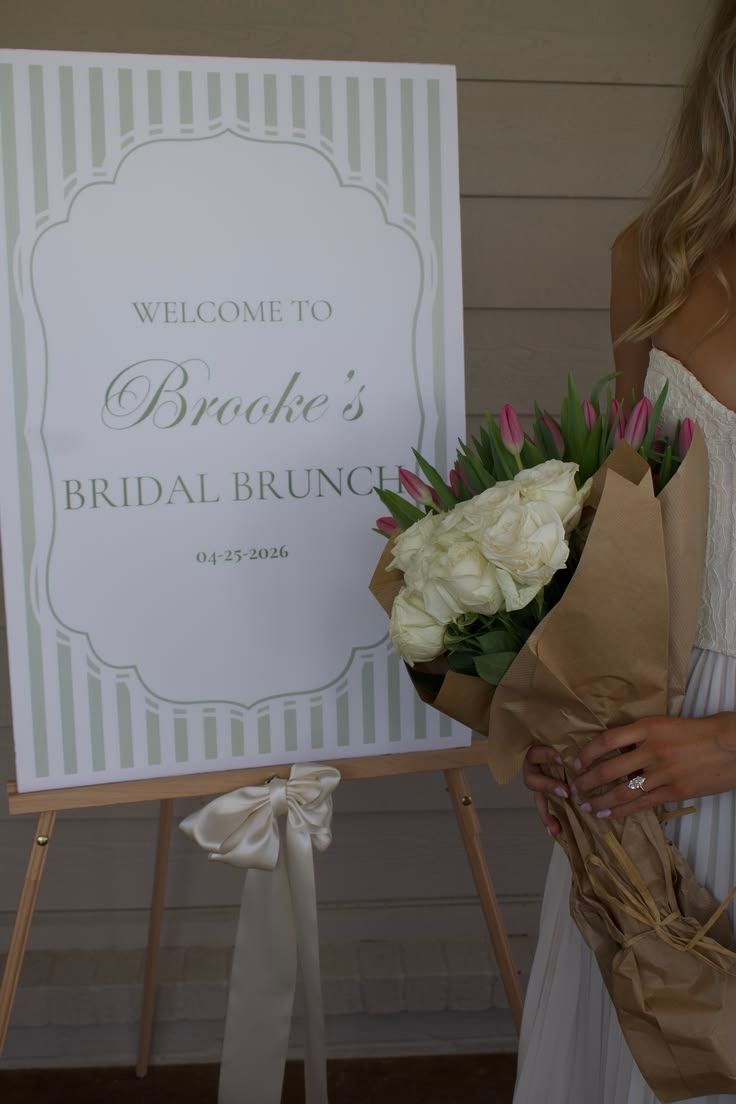 Brooke's Bridal Brunch welcome sign on easel with white bow, beside bride holding white roses bouquet.
