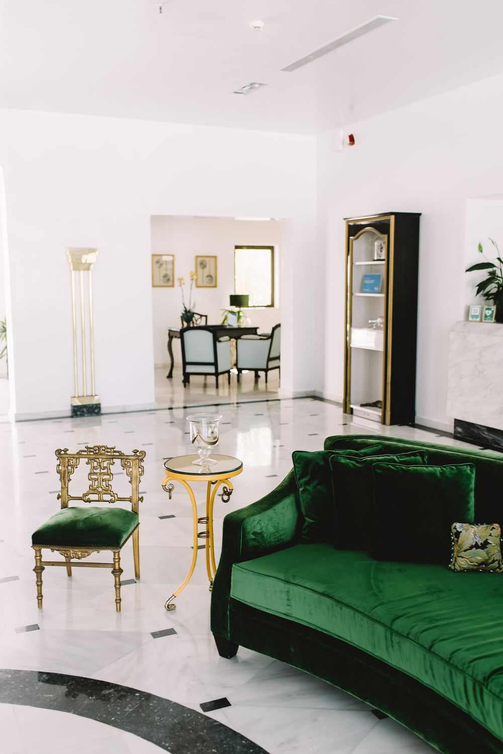 Elegant lobby with green velvet sofa, ornate gold chair, marble floors, and classic black display cabinet.