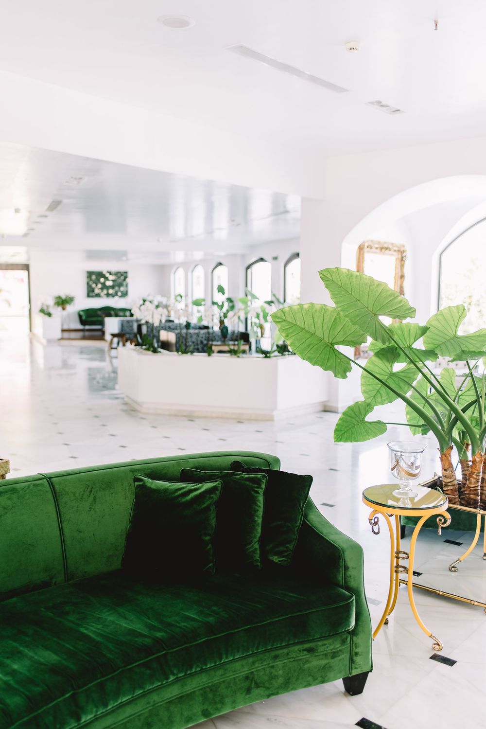 Elegant hotel lobby with green velvet sofa, tropical plants, gold side table, and white marble floors.