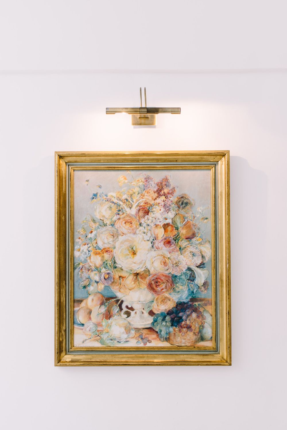 Gold framed floral painting illuminated by a brass picture light on a white wall.
