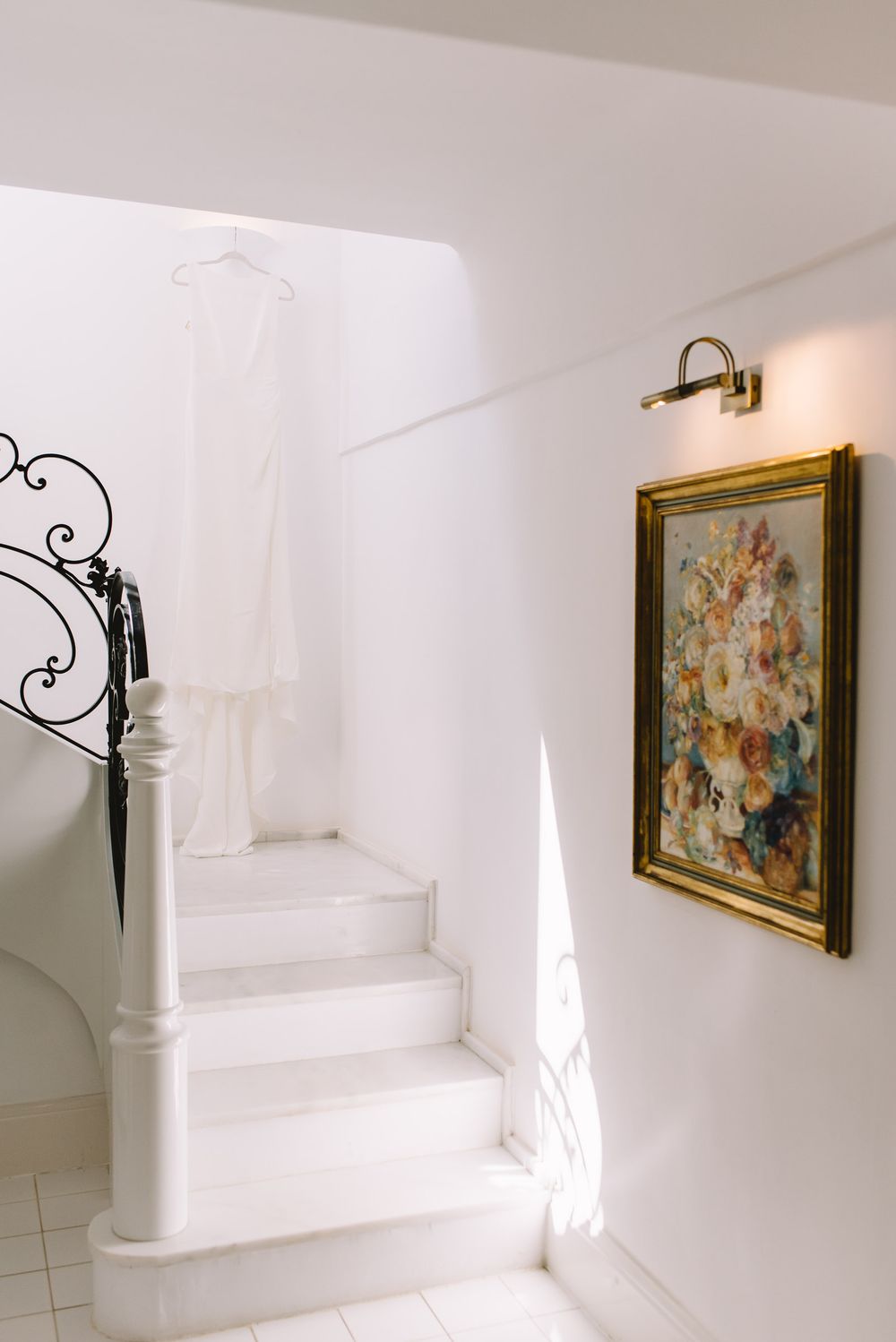 White wedding dress hanging on ornate staircase railing beside floral painting in elegant white interior.