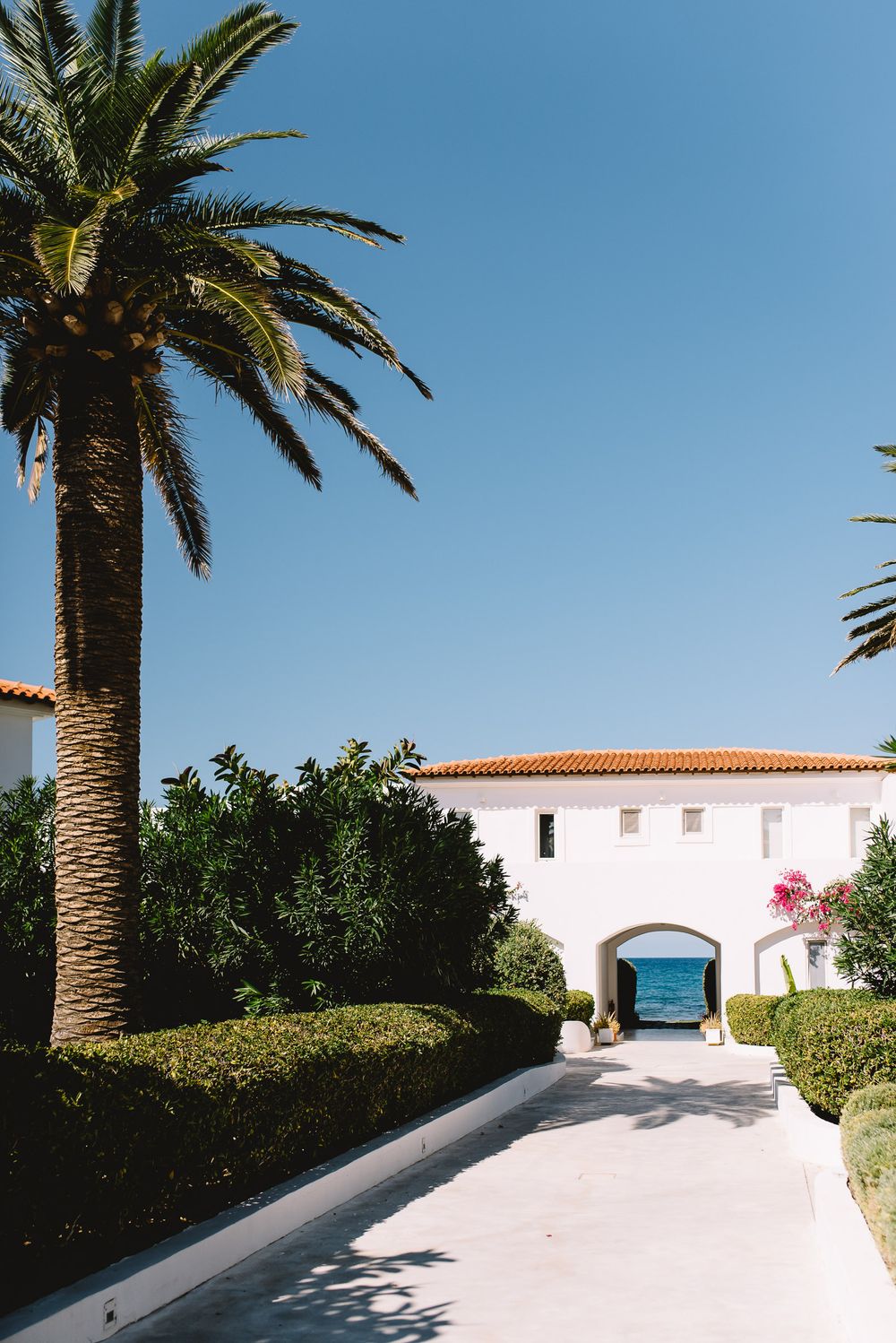 White Mediterranean villa with red-tile roof, palm tree, and ocean view through archway on sunny day.