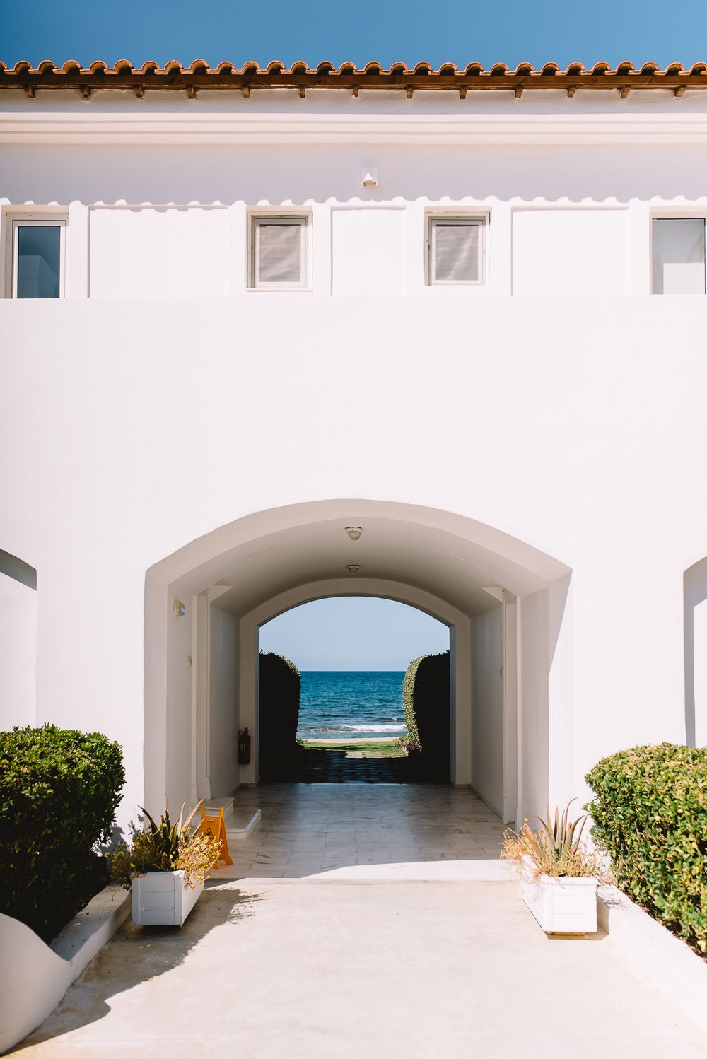 White Mediterranean arch passageway leading to stunning blue ocean view, flanked by trimmed hedges and terracotta roof.