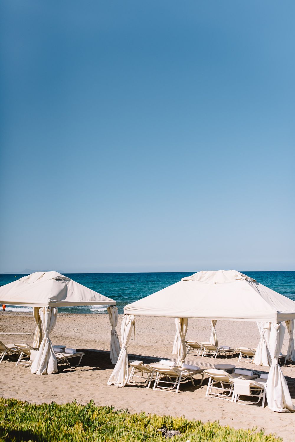 White beach cabanas with lounge chairs on a sandy shore, overlooking a calm turquoise sea under a clear blue sky.