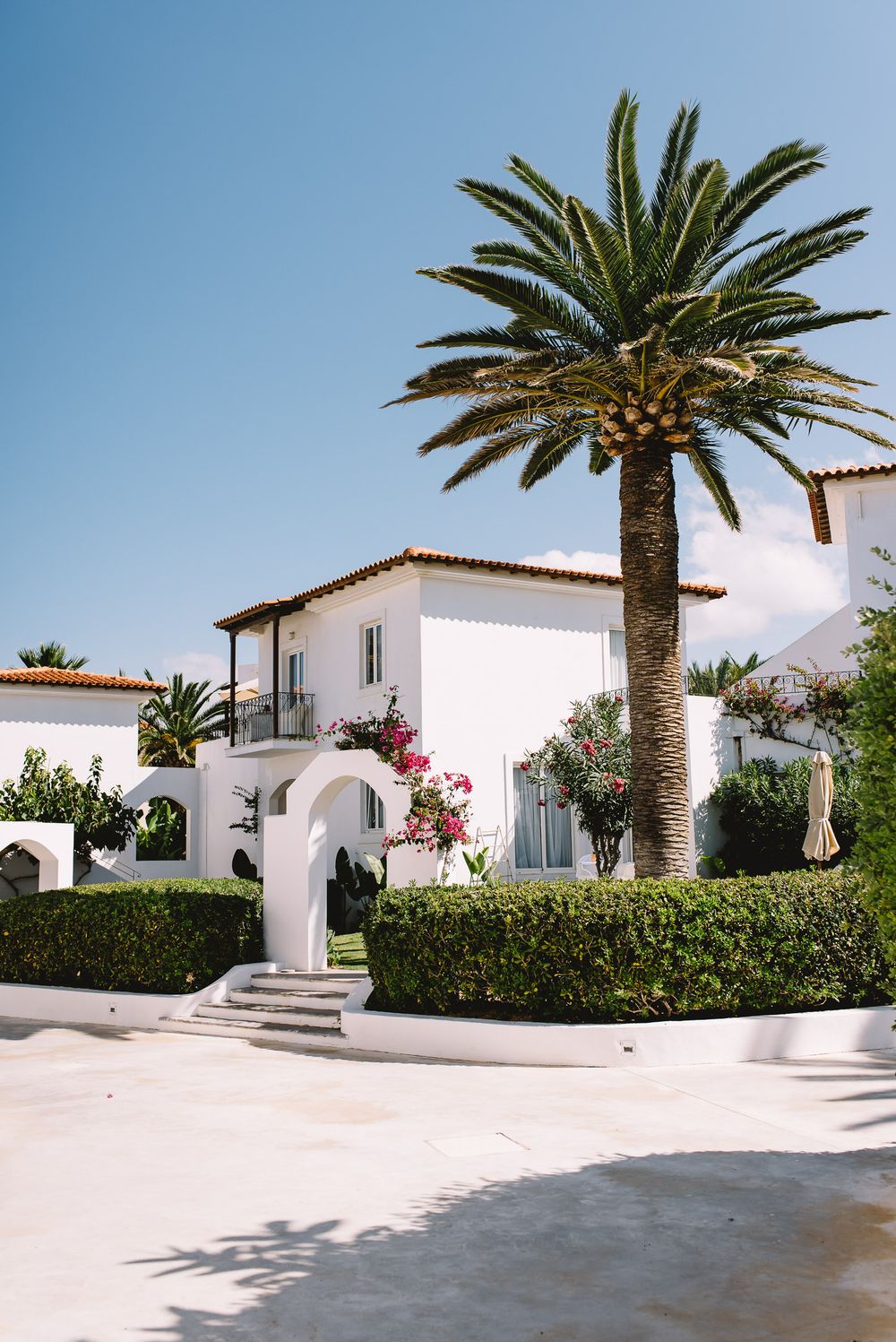 White Mediterranean villa with palm tree, bougainvillea, and manicured hedges under a clear blue sky.