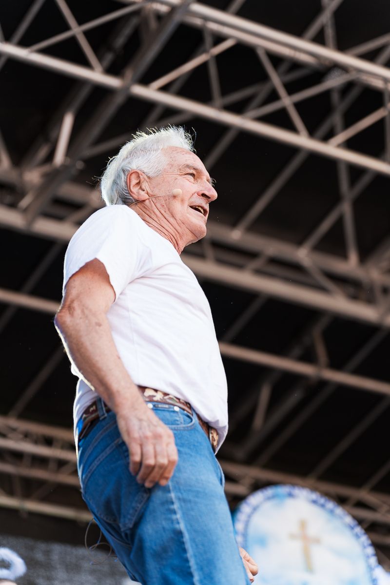 Elderly man in white t-shirt and jeans performing on outdoor stage with metal roof structure visible above.