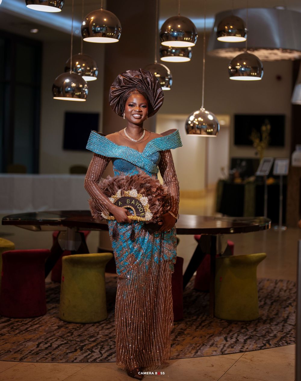 Woman in elegant African traditional attire with headwrap and fan poses under metallic pendant lights in modern venue.