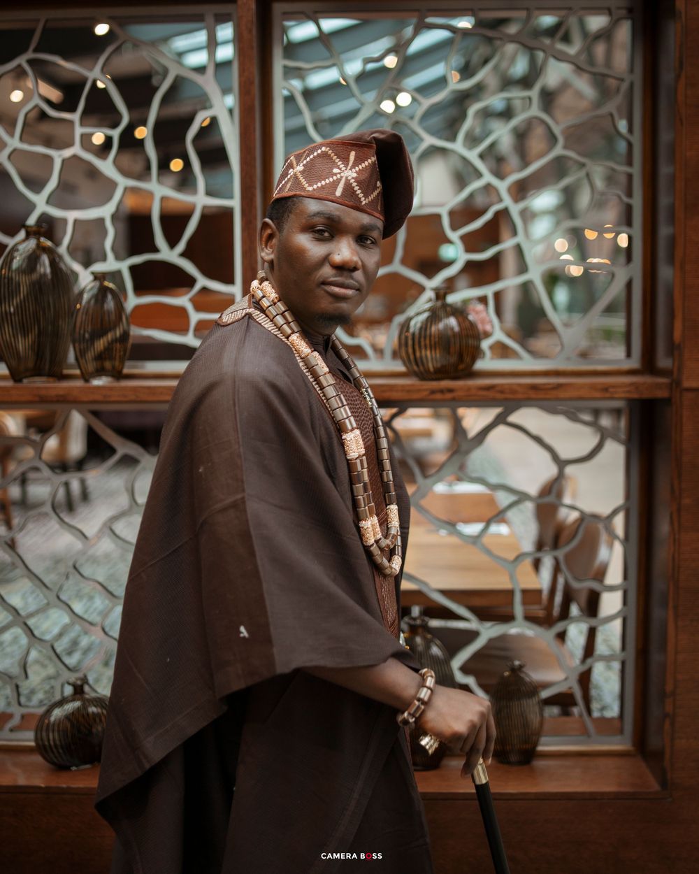 Man in traditional African attire with beaded necklace, cap, and walking stick poses before decorative wooden window.