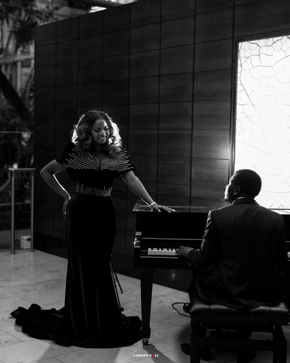 Black and white photo of a woman in an elegant gown standing by a piano while a man plays in a formal setting.