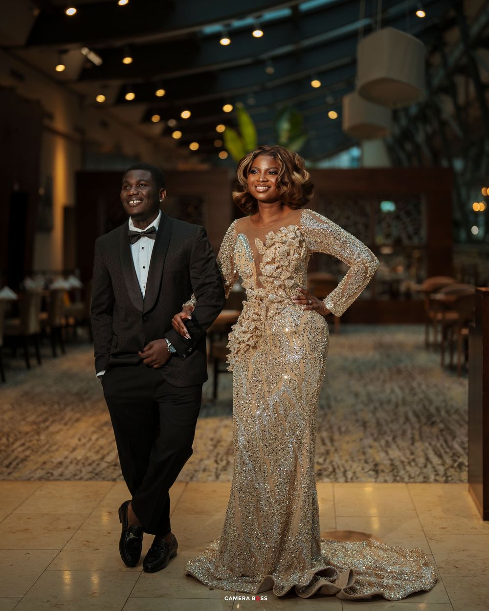 Elegant couple posing in formal attire, man in black tuxedo and woman in gold sequined gown, at upscale venue.