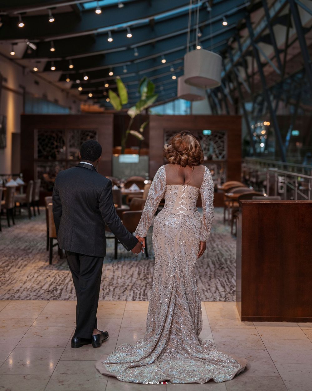 Couple holding hands in elegant venue, woman in sequined gown with train, man in dark suit, romantic wedding photo.