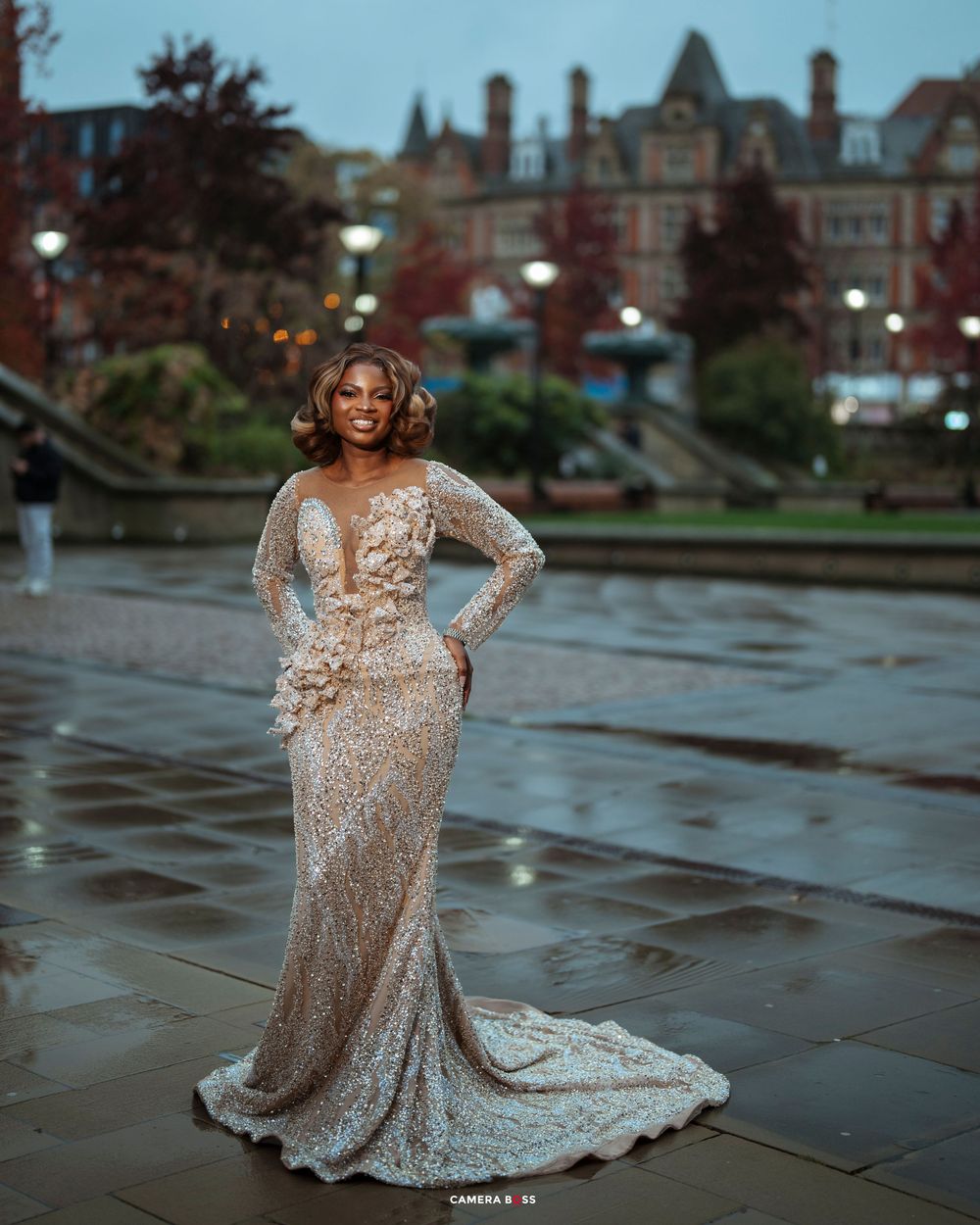 Woman in elegant sequined mermaid gown with floral details poses outdoors near historic building.