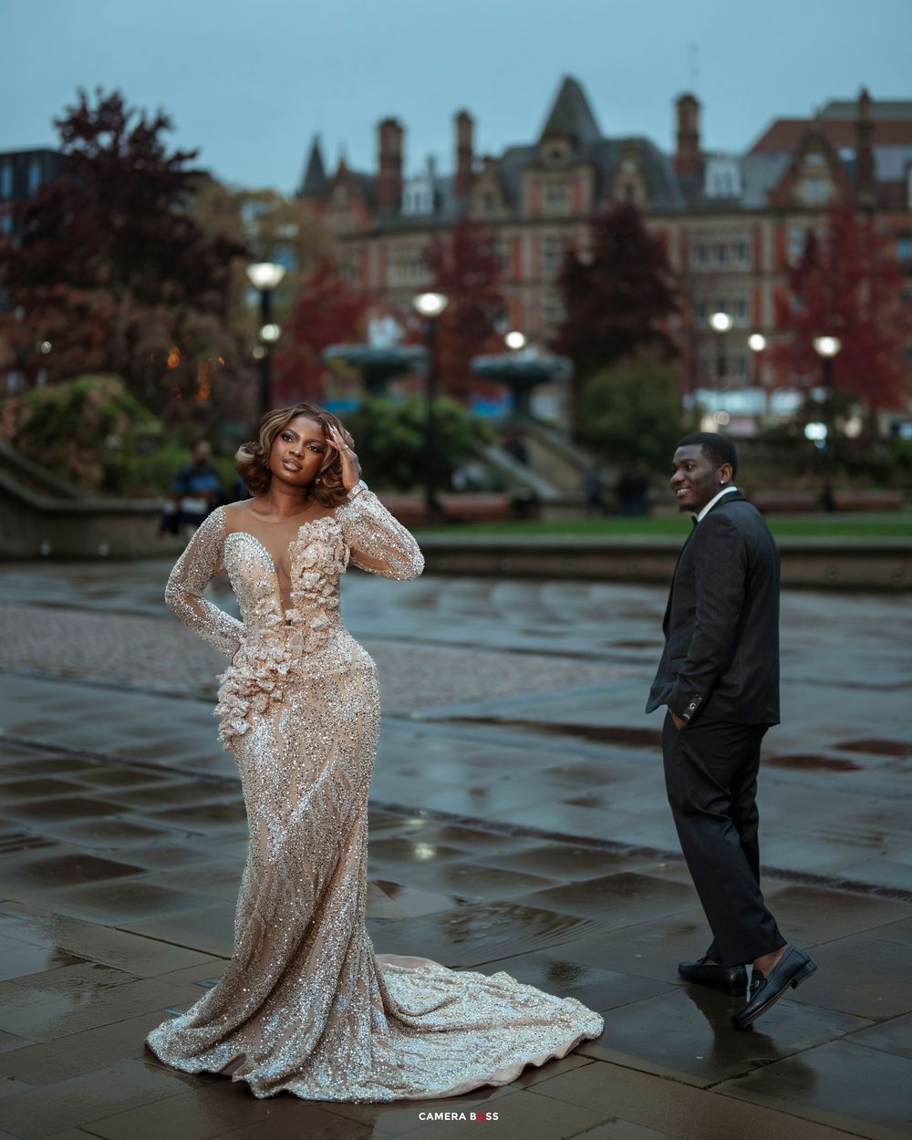 Bride in stunning sequined mermaid gown poses with groom in suit at rainy outdoor plaza with historic building backdrop.