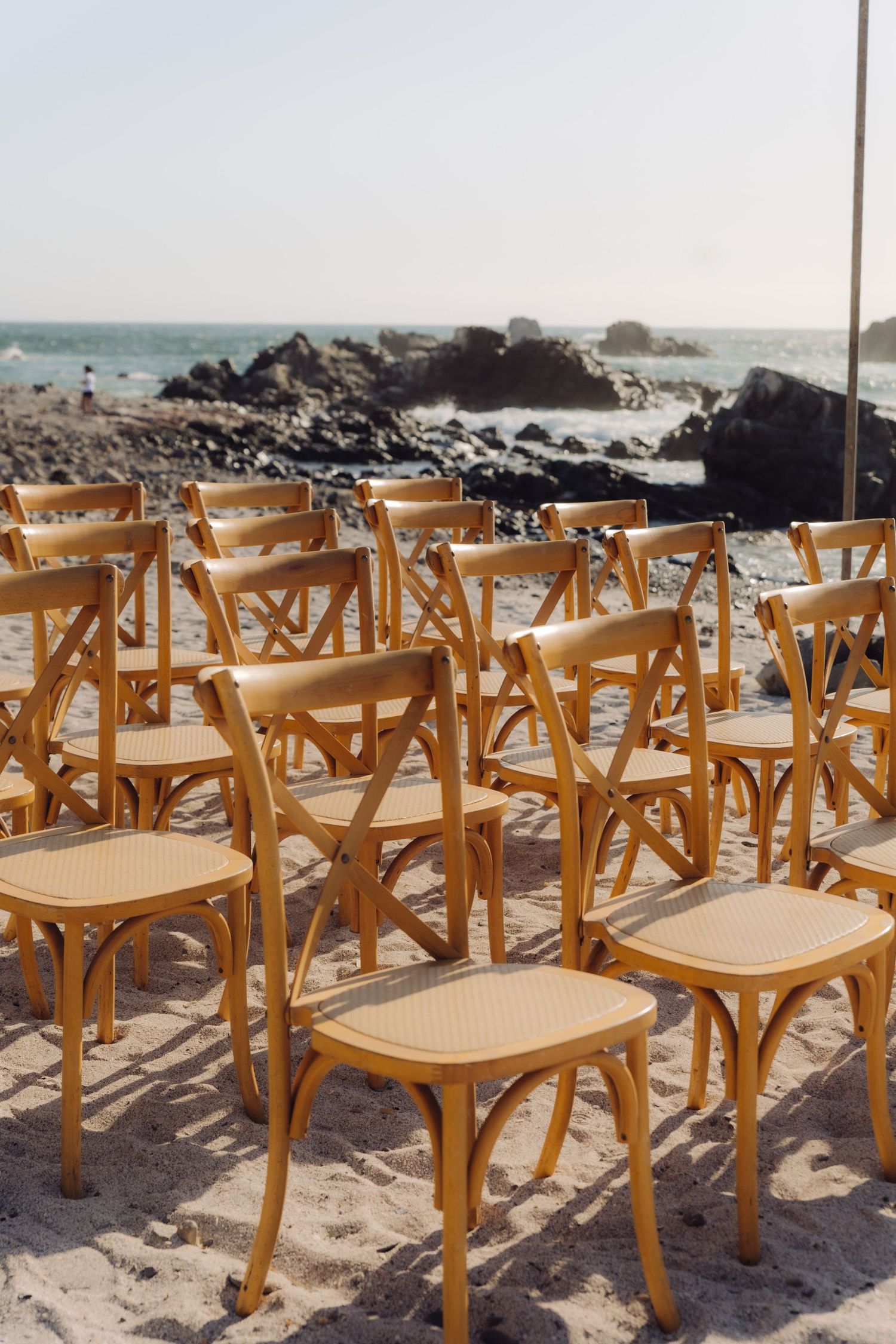 Rows of wooden cross-back chairs arranged on sandy beach for an outdoor wedding ceremony.