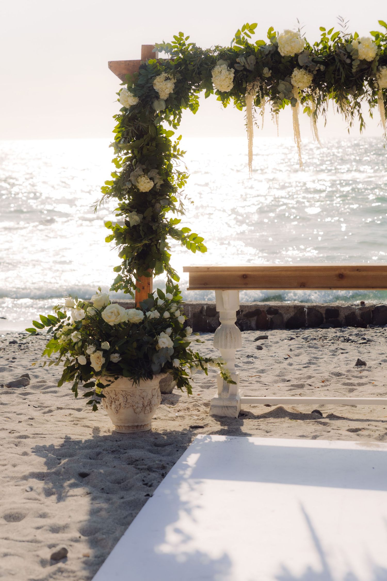 White floral arrangement in vintage vase beside a wooden wedding arch decorated with greenery by the sea.
