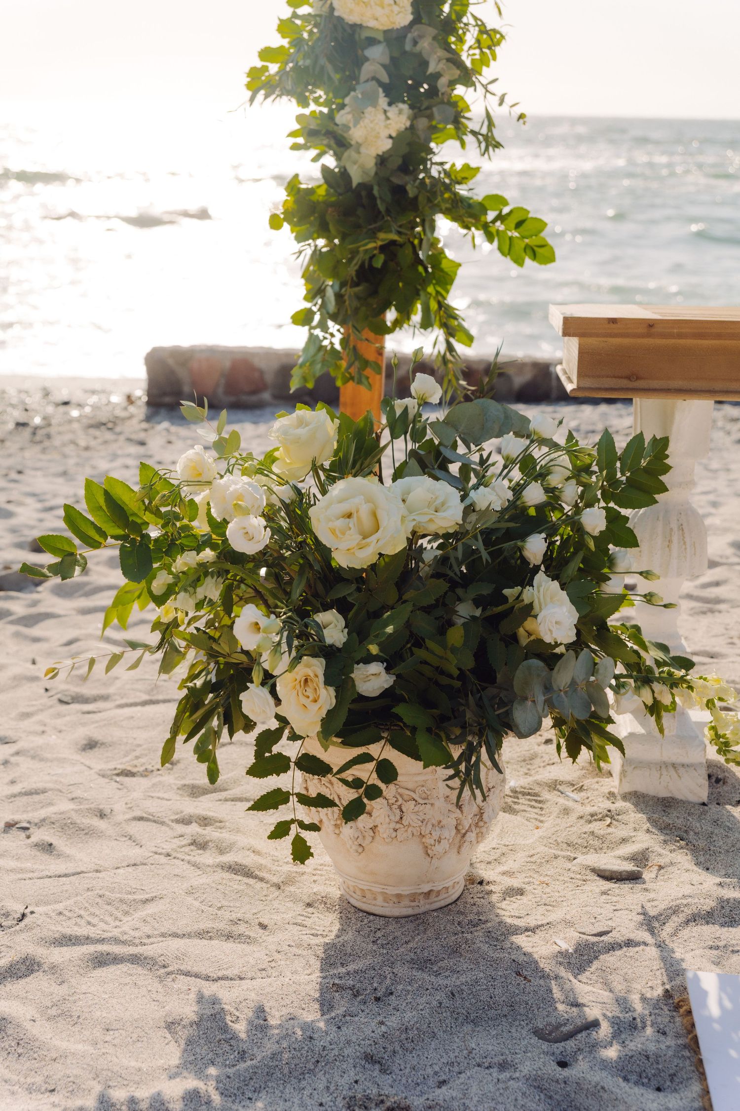 Elegant white roses and greenery bouquet in rustic vase on sandy beach wedding ceremony site.
