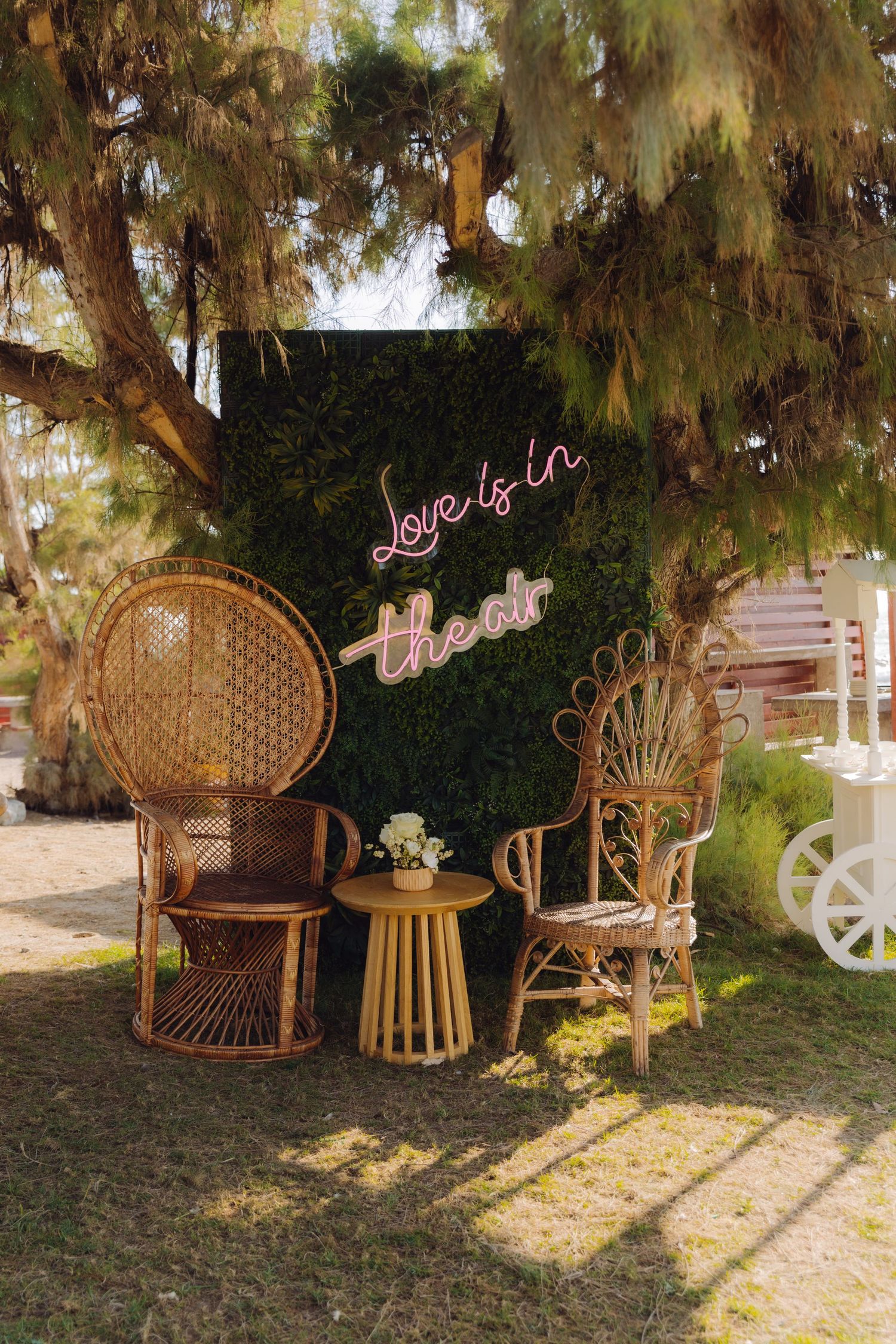 Boho wedding photo backdrop with rattan peacock chairs and neon Love Is In The Air sign.