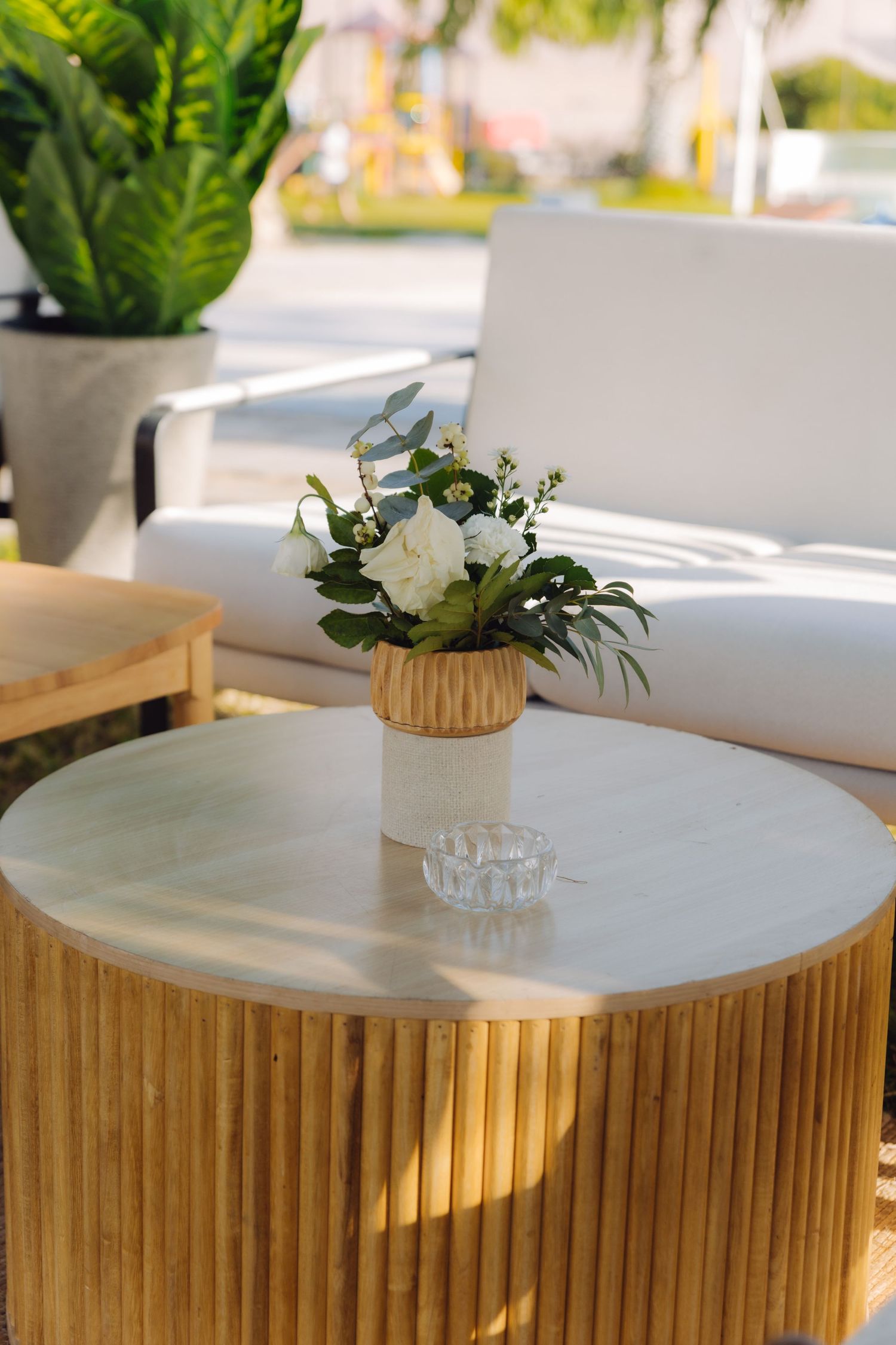 Elegant outdoor lounge with wooden table featuring white flowers and glass ashtray beside white sofa.