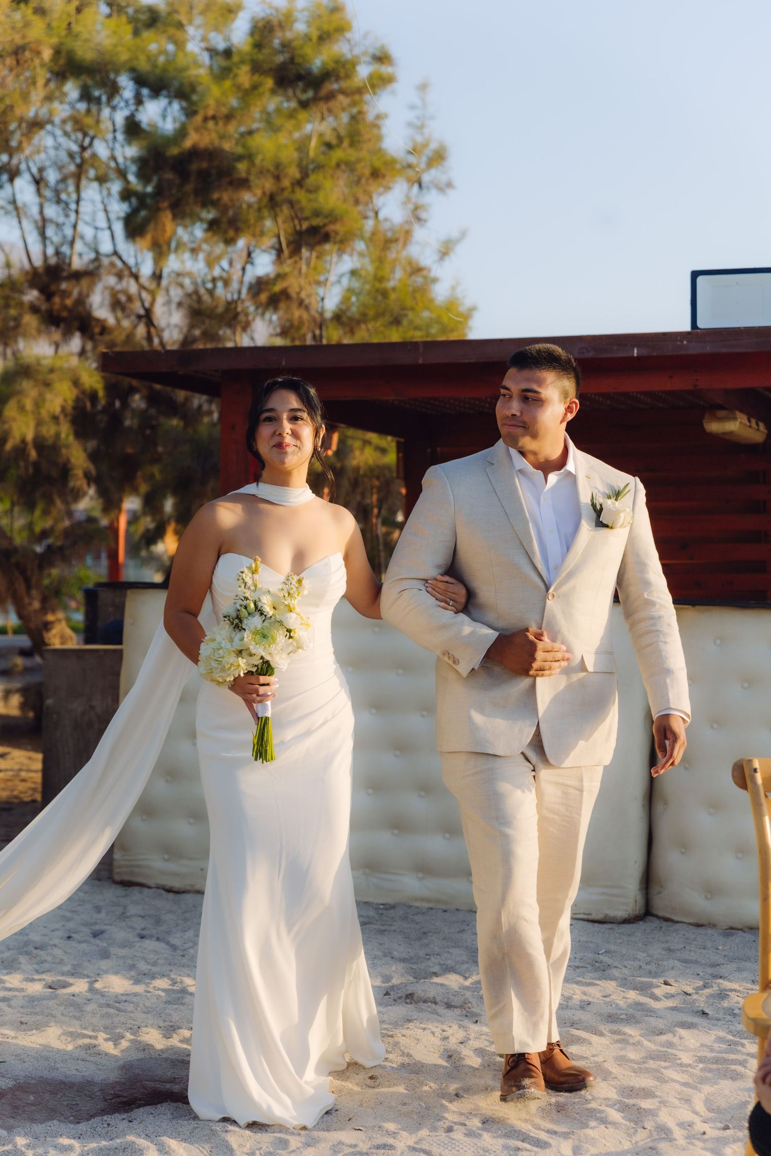 Bride in white halter gown holding bouquet walks with groom in beige suit on sandy beach.