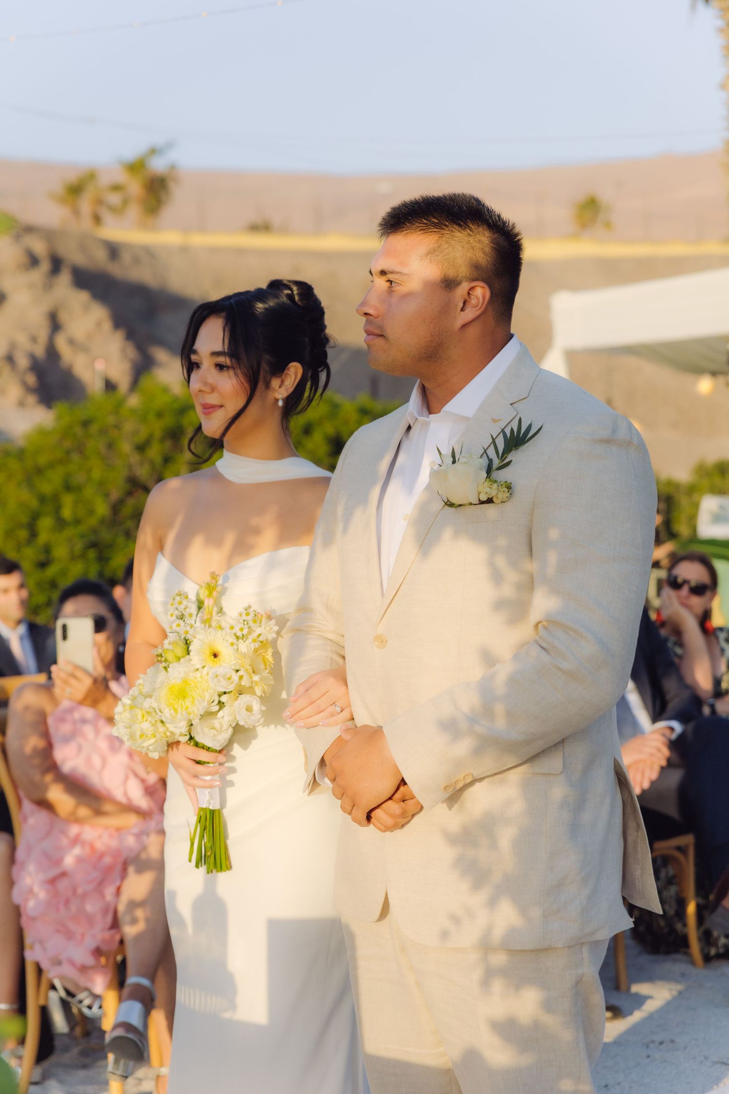 Bride holding white bouquet stands with groom in cream suit during outdoor wedding ceremony.