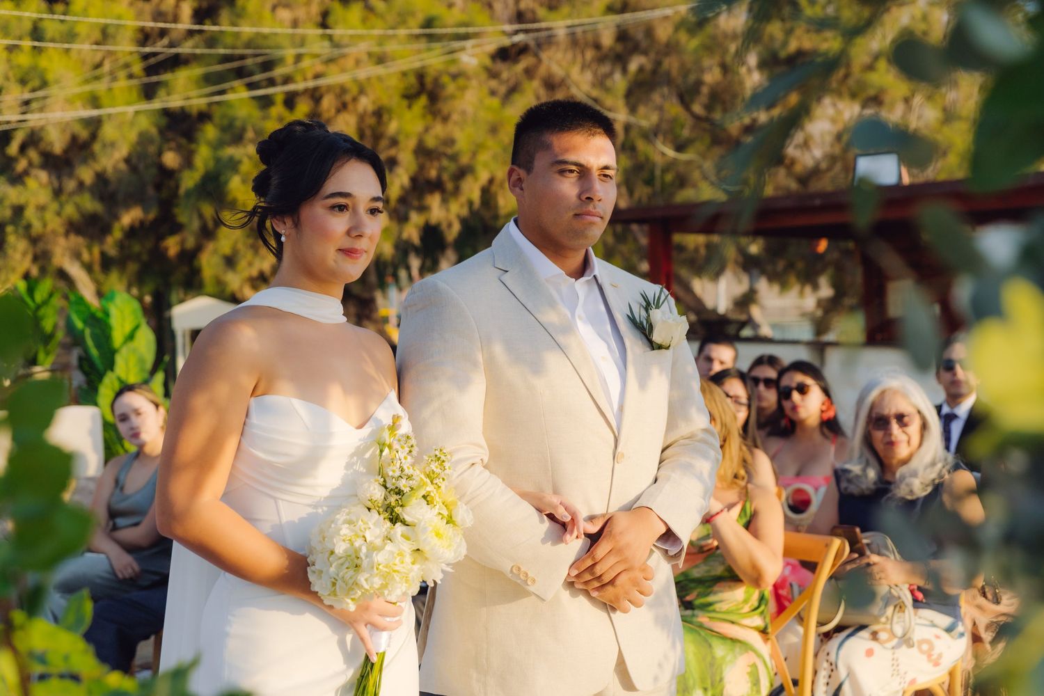 Bride in strapless white gown and groom in beige suit stand at outdoor wedding ceremony altar.