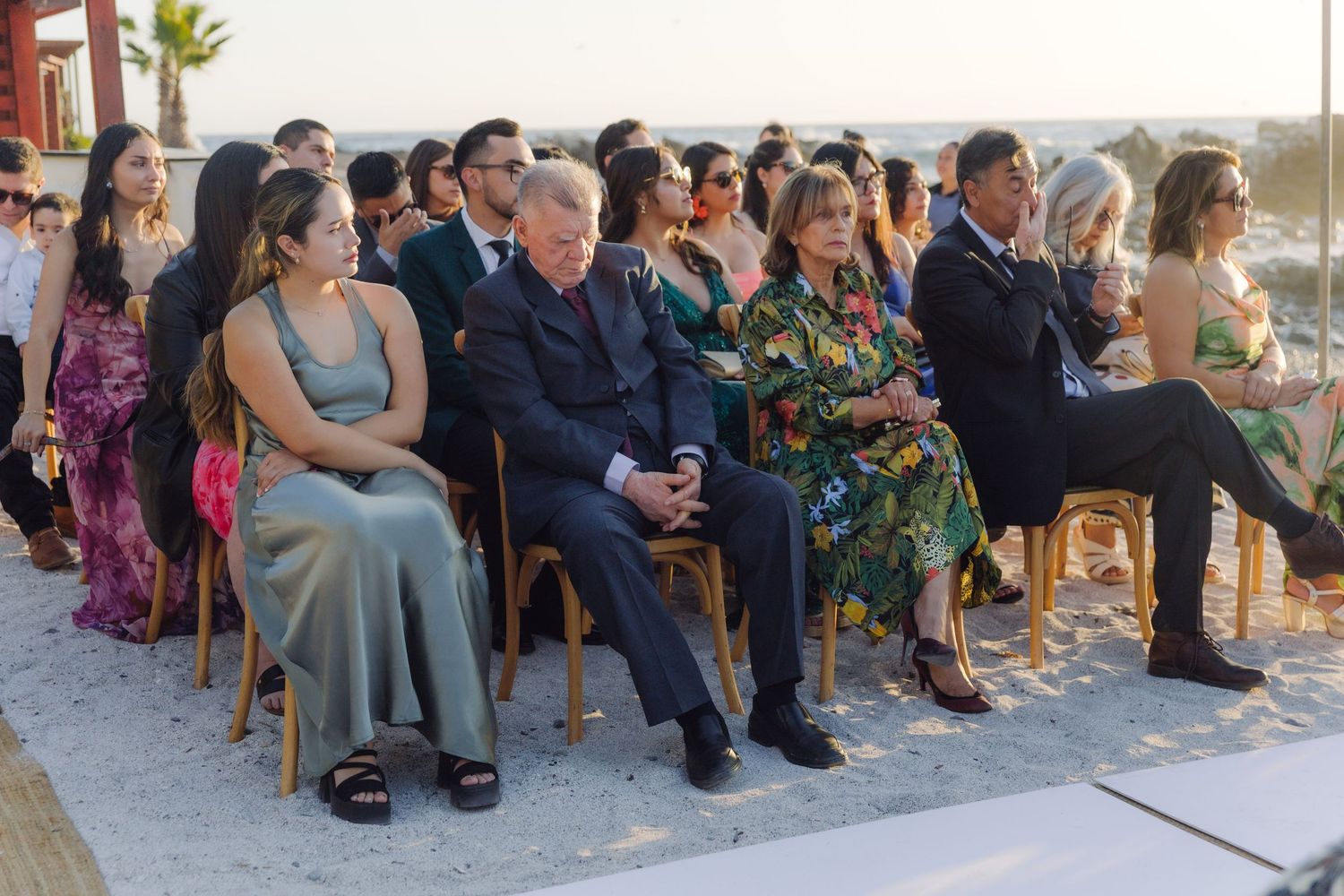 Wedding guests seated in wooden chairs watching outdoor beach ceremony in evening sunlight.