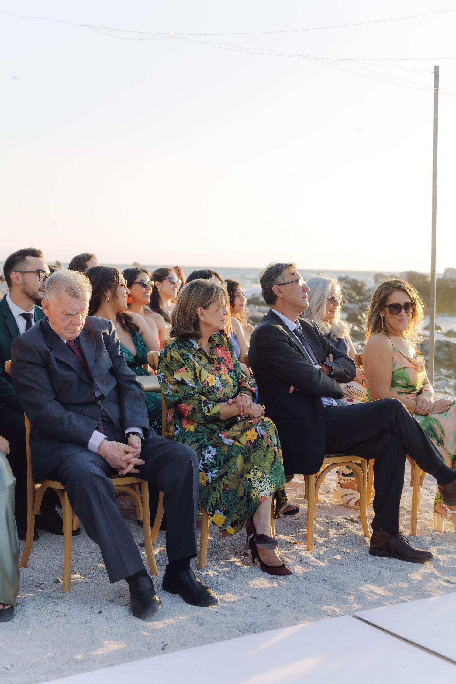 Guests seated on wooden chairs at an outdoor beach wedding ceremony during golden hour.