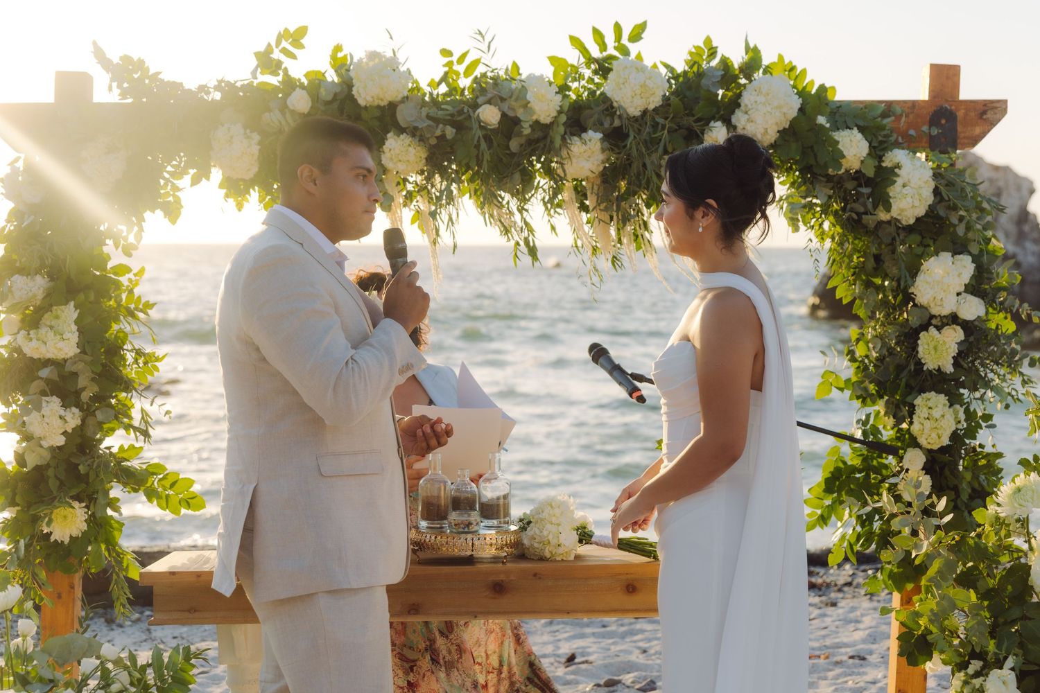 Groom reading vows to bride under a white floral arch at a beach wedding ceremony.