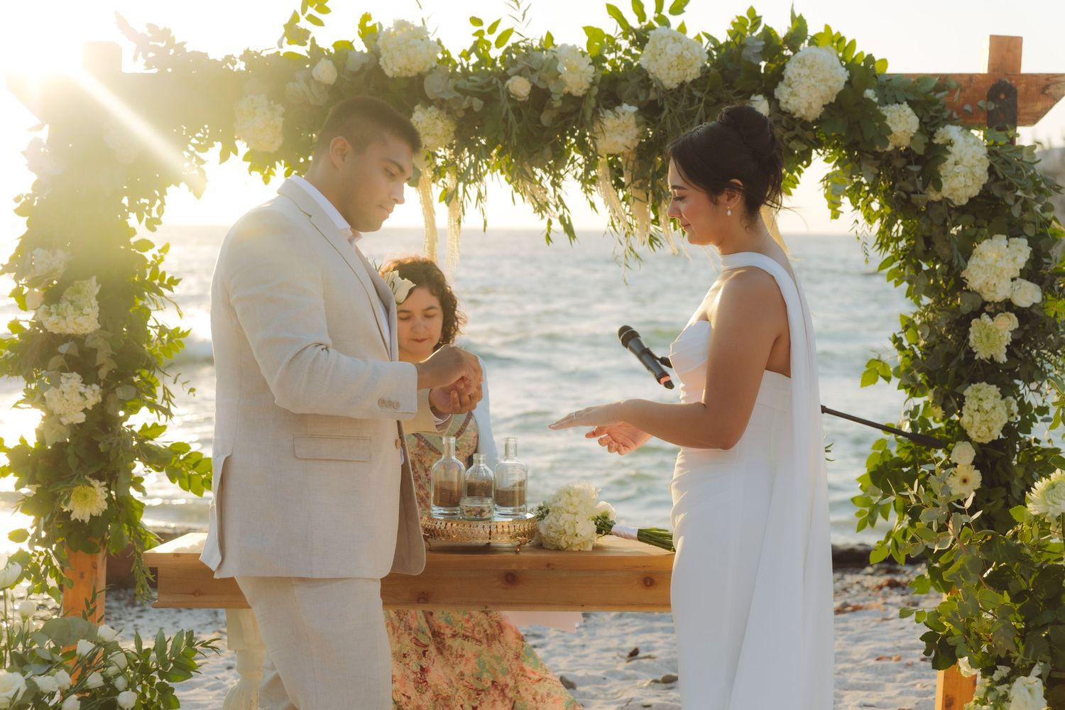 Bride reading vows to groom under a lush floral arch at a sunset beach wedding.