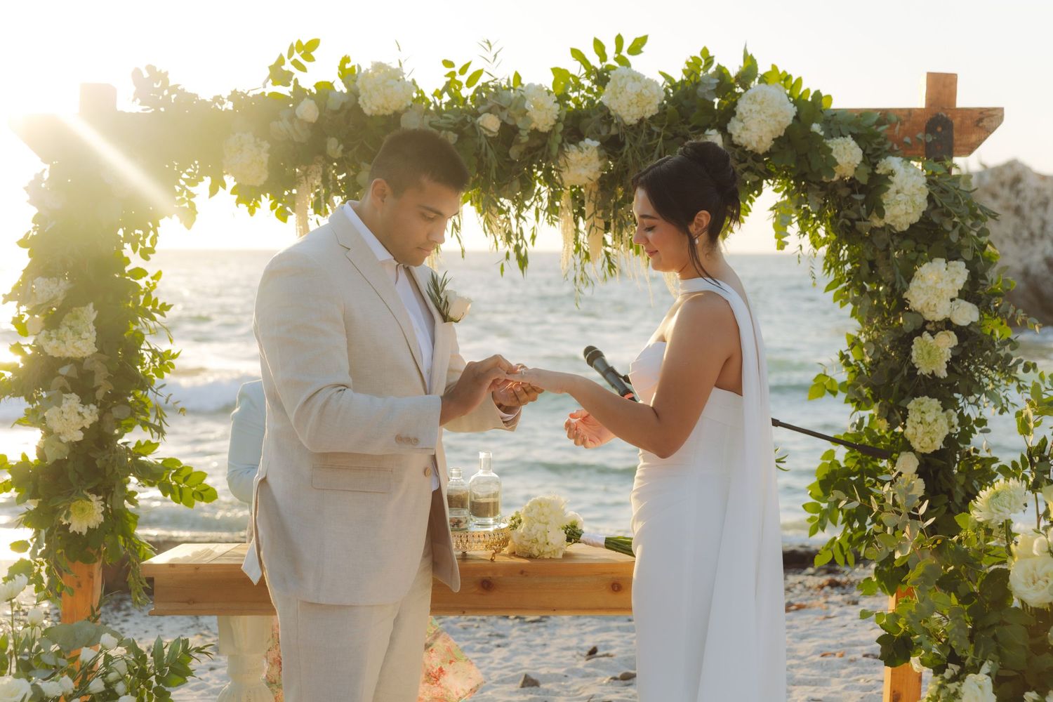 Groom placing wedding ring on brides finger under a floral arch at beach ceremony.