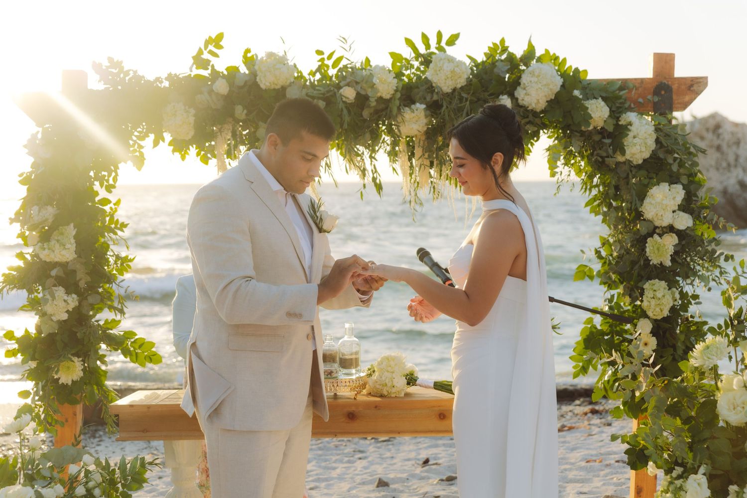 Couple exchanging rings under a white floral arch at a golden hour beach wedding.