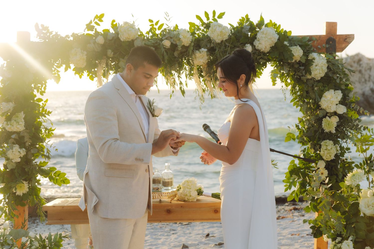 Groom sliding ring onto brides finger under a flower-adorned arch at beach wedding.