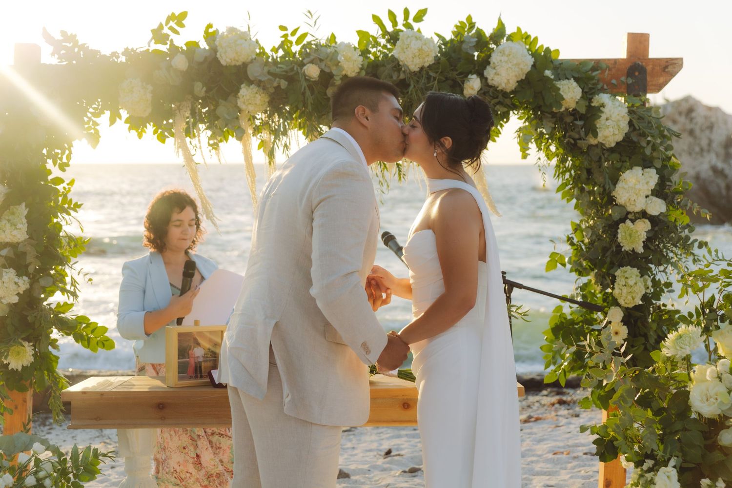 Newlyweds sharing first kiss under a floral arch at a romantic sunset beach wedding.