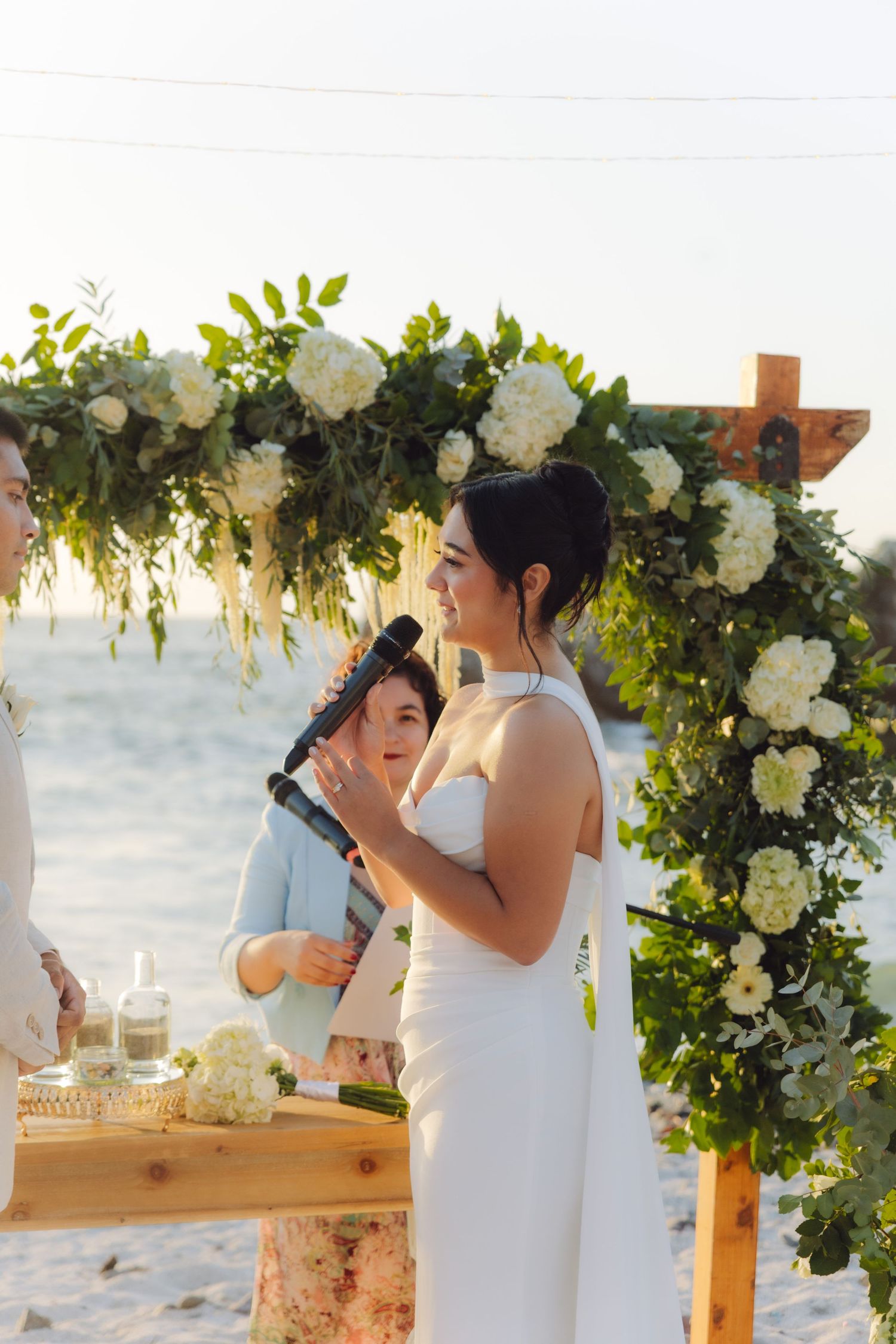 Smiling bride holding a scroll at a beach wedding ceremony under a white floral arch.