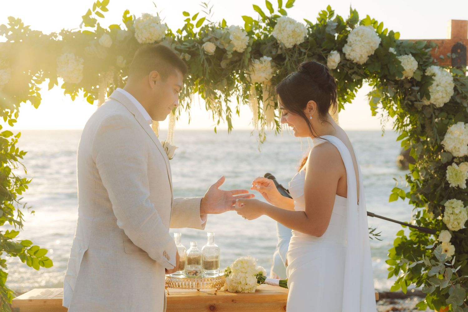 Couple exchanging rings at oceanside wedding ceremony under floral arch with white blooms at sunset.