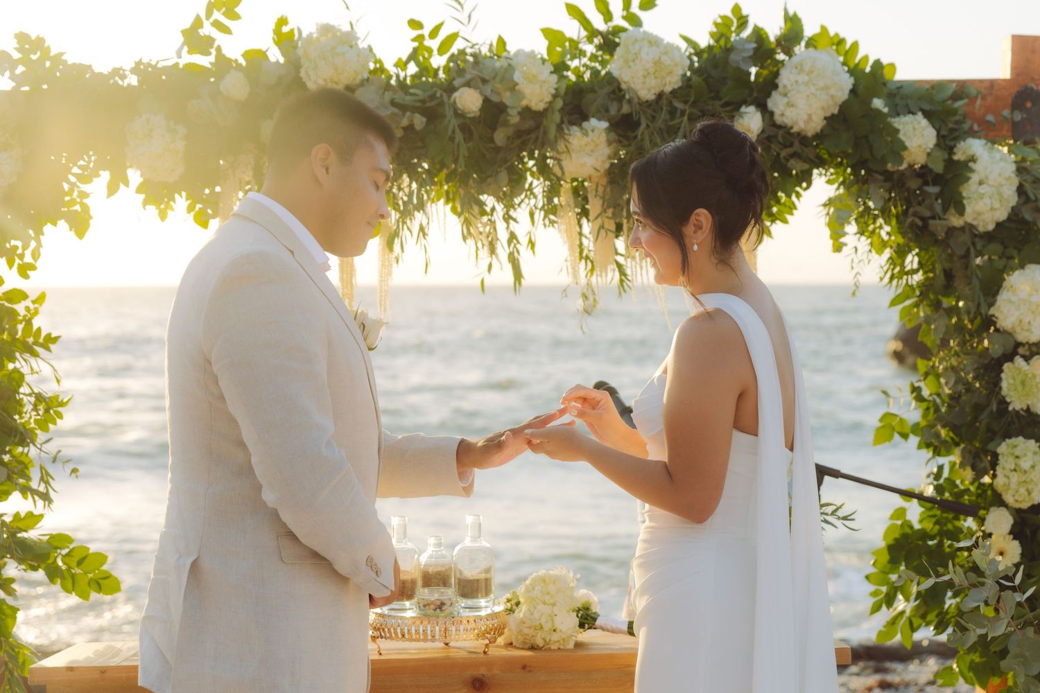 Bride and groom holding hands during ring exchange at beach wedding under lush greenery floral arch.