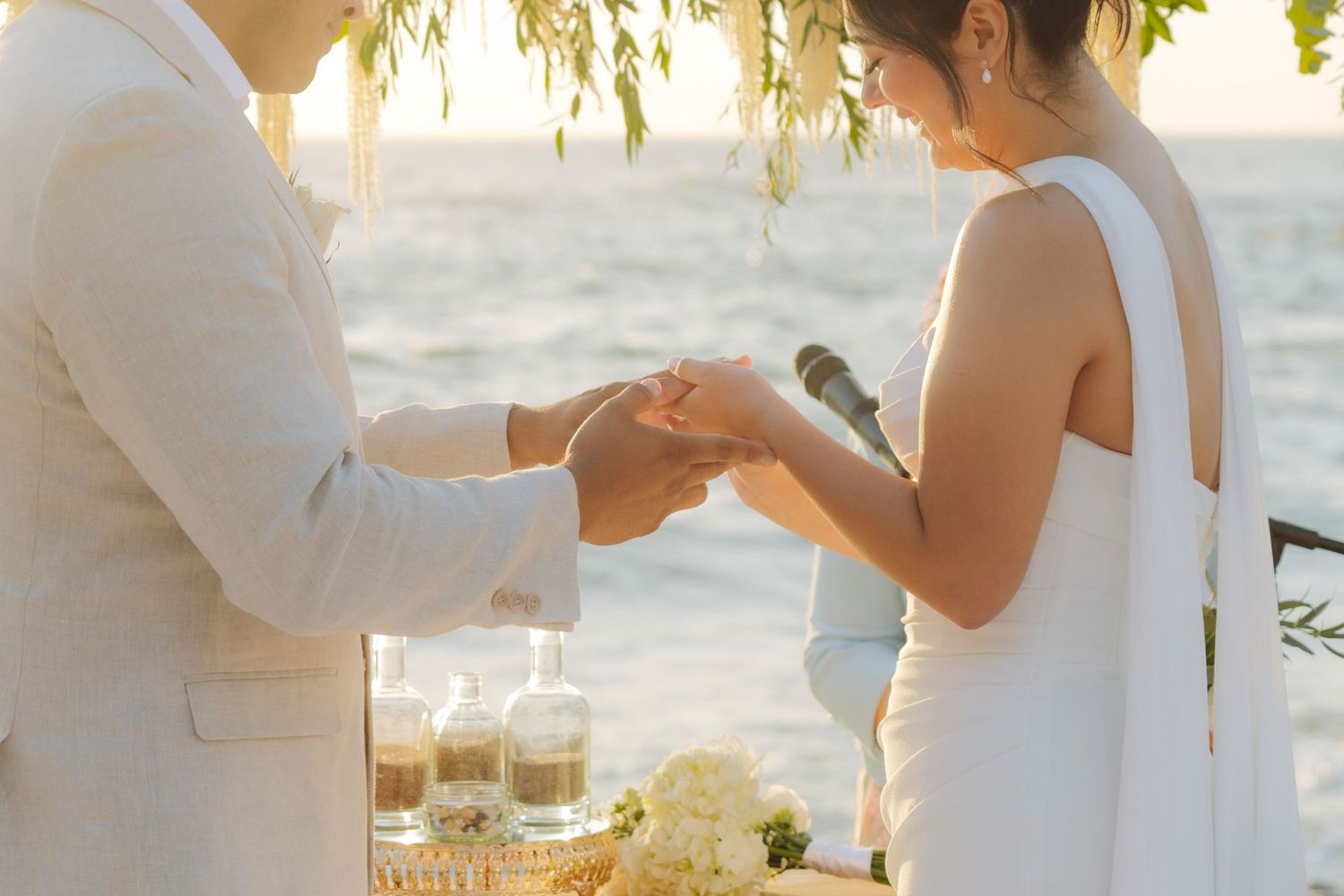 Close-up of bride placing wedding ring on grooms hand at seaside ceremony with sand bottles nearby.