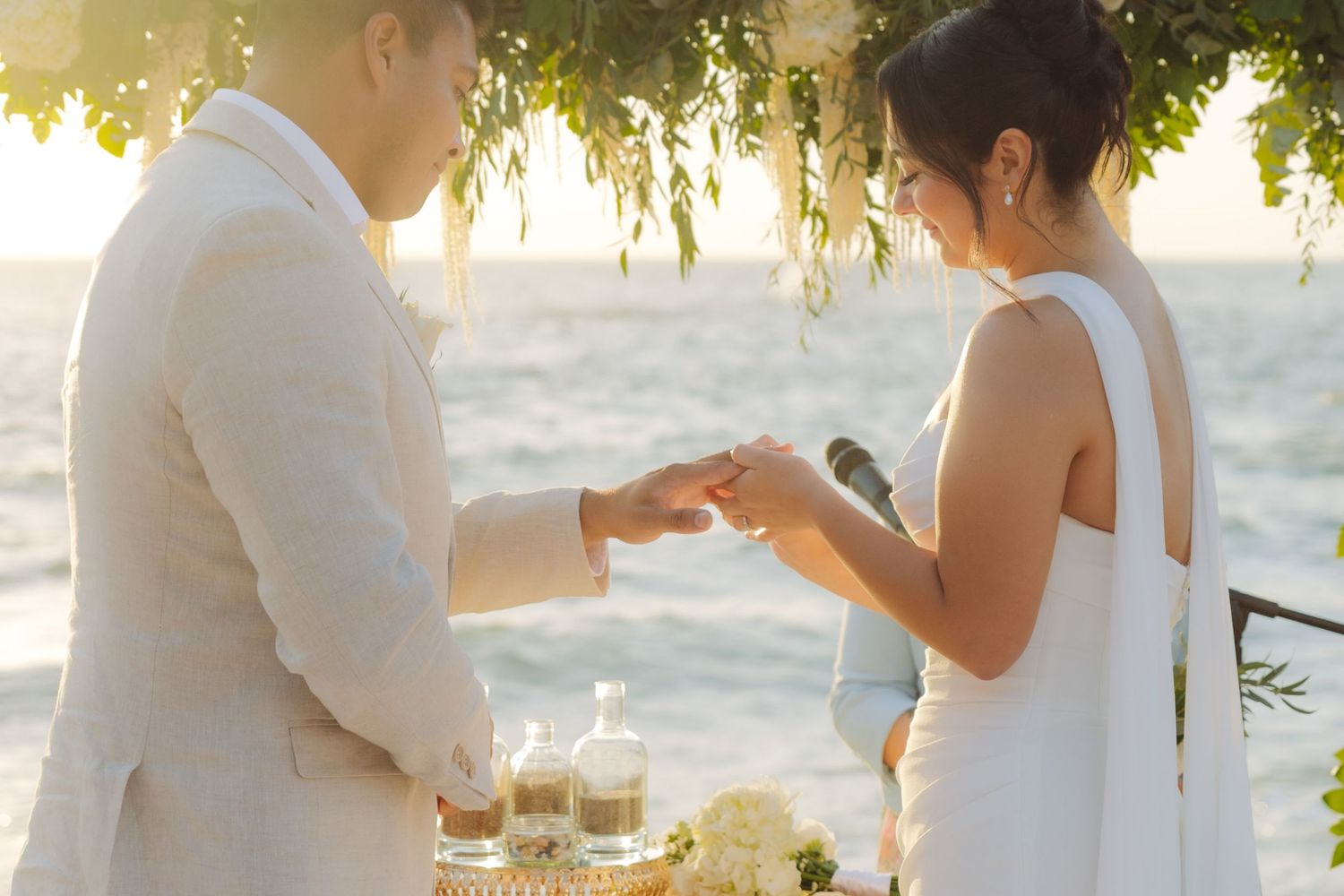 Bride sliding wedding ring onto grooms finger during outdoor beach ceremony at golden sunset.
