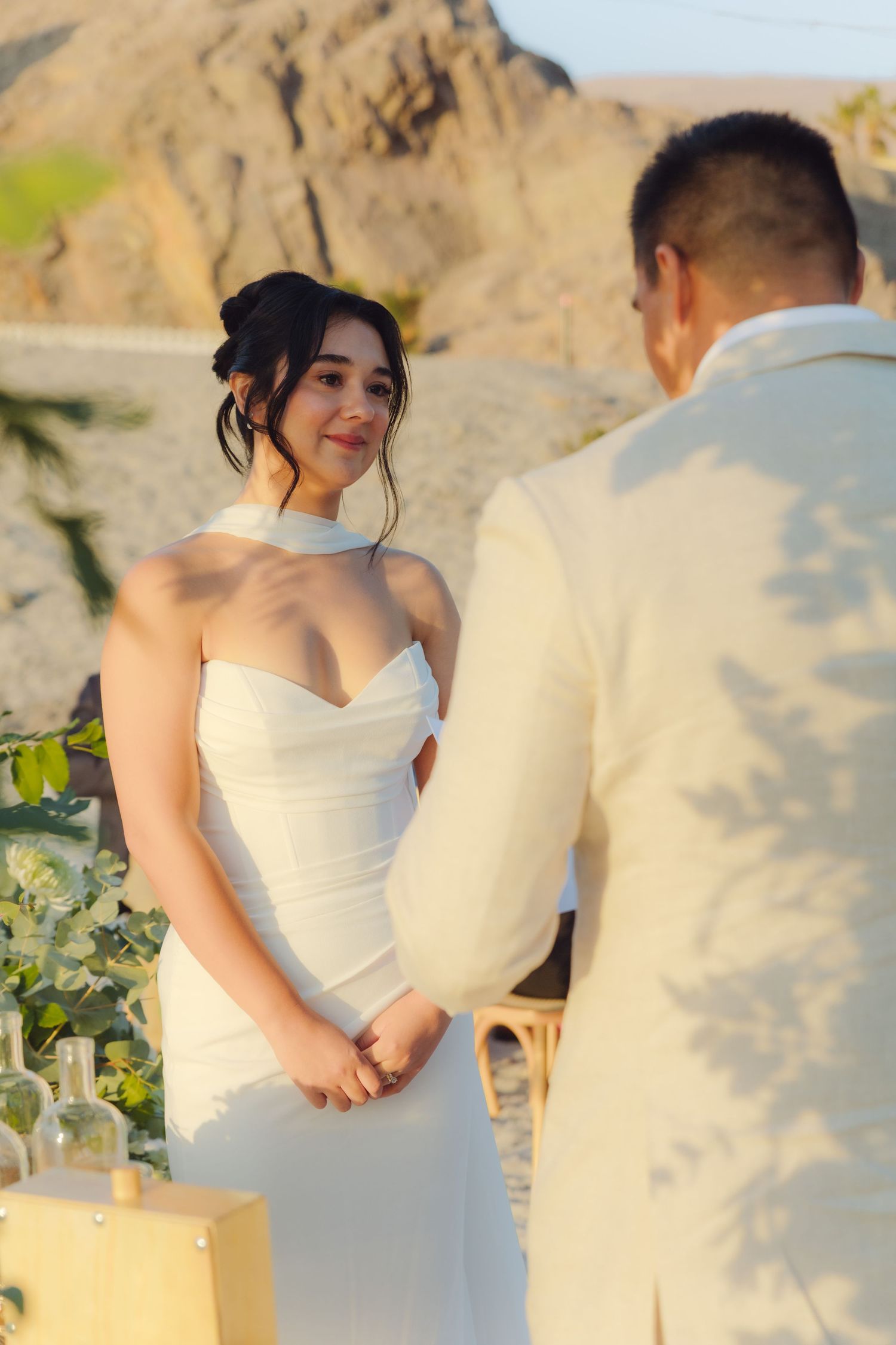 Smiling bride in halter wedding gown listens to groom recite vows at desert outdoor ceremony.