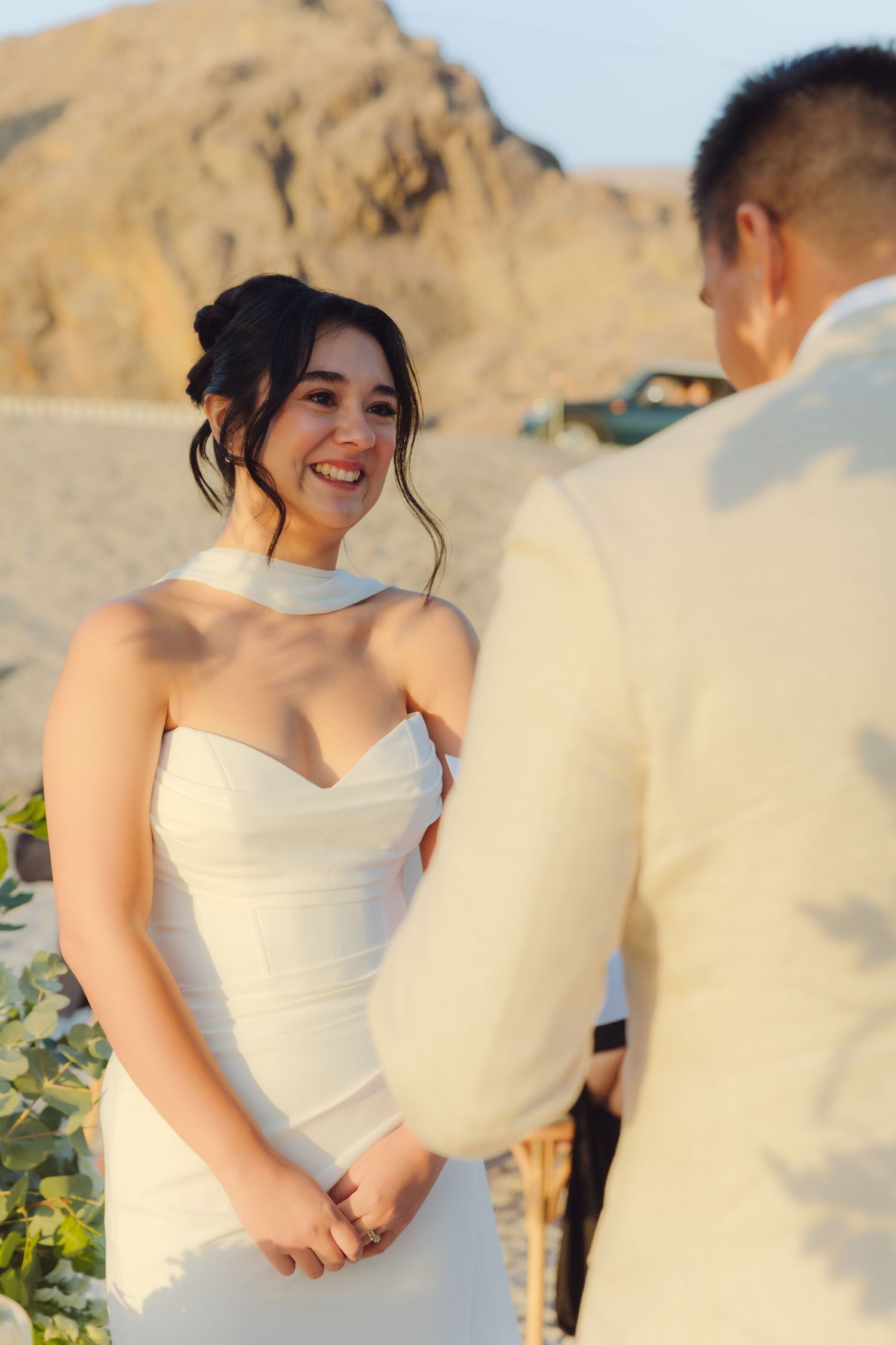 Radiant bride in white halter dress smiles joyfully at groom during outdoor desert wedding ceremony.