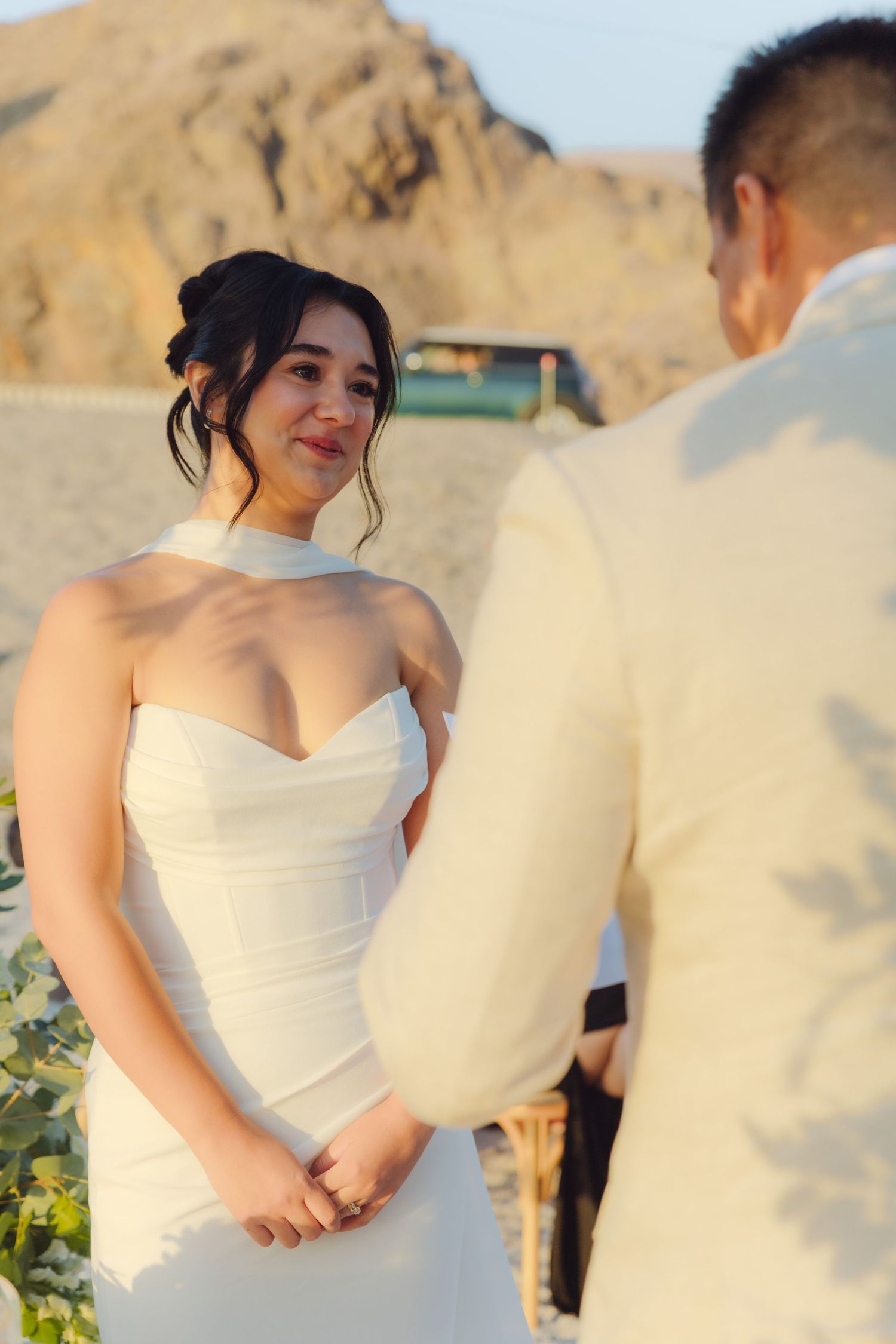 Emotional bride in elegant white halter gown listens to grooms vows at scenic desert wedding.