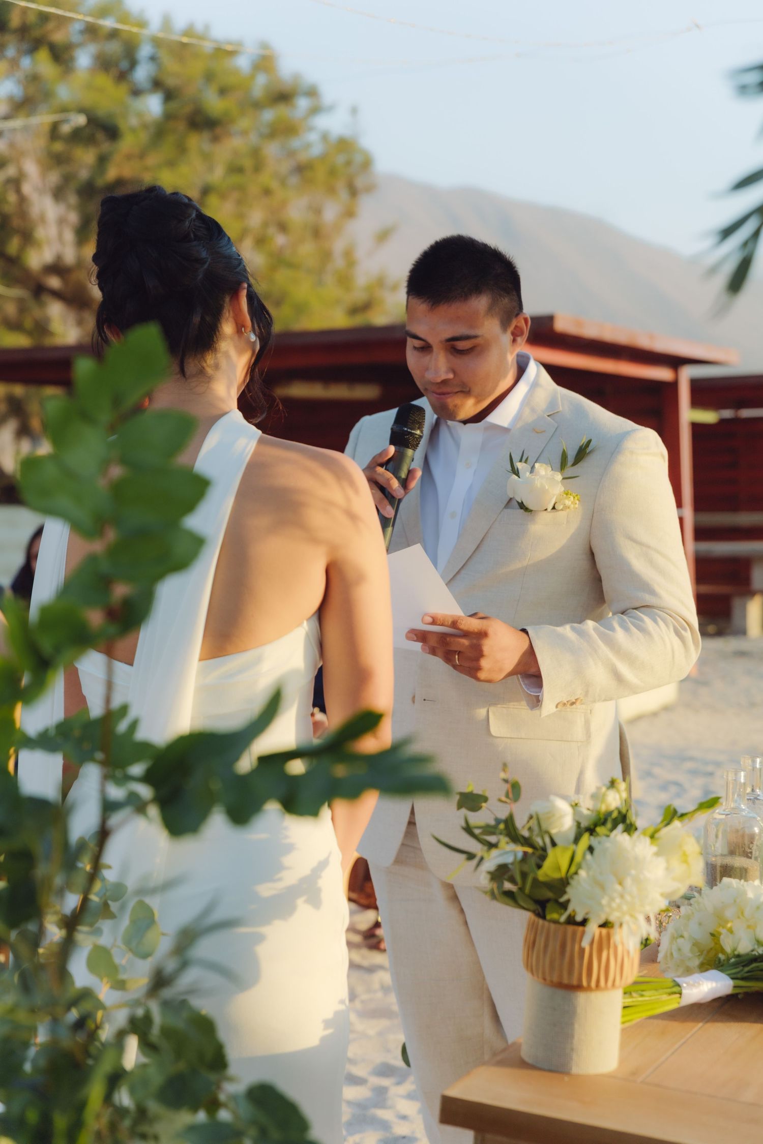 Groom in cream suit reads wedding vows into microphone at outdoor beach ceremony at sunset.