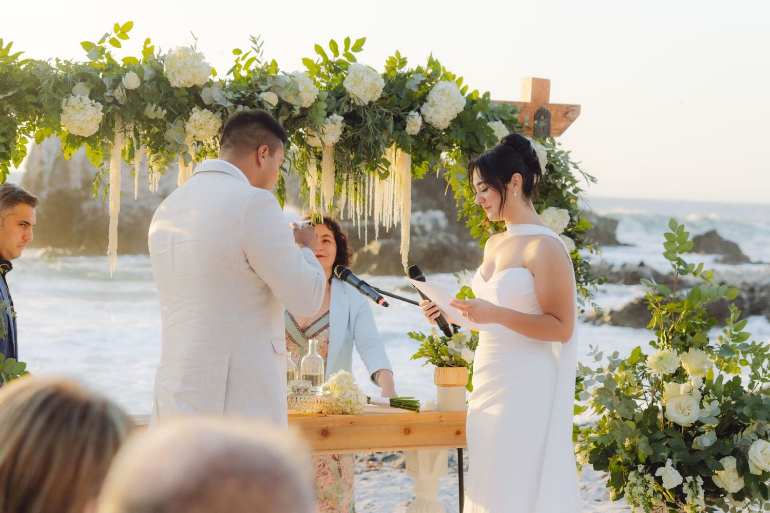 Bride and groom exchange vows under lush white floral arch at oceanside wedding ceremony.