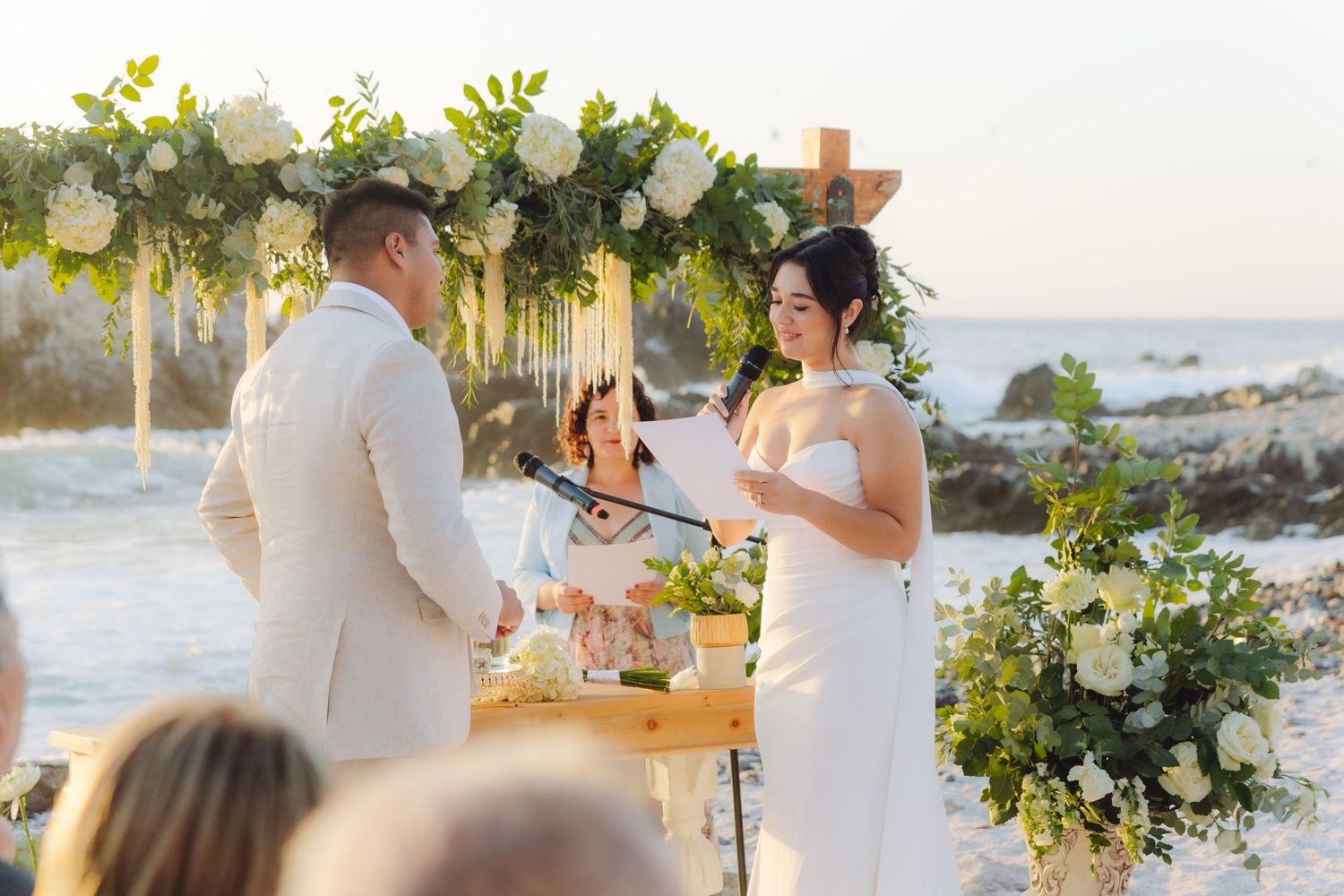 Bride reads vows to groom under elegant white floral arch at beautiful beachside wedding ceremony.
