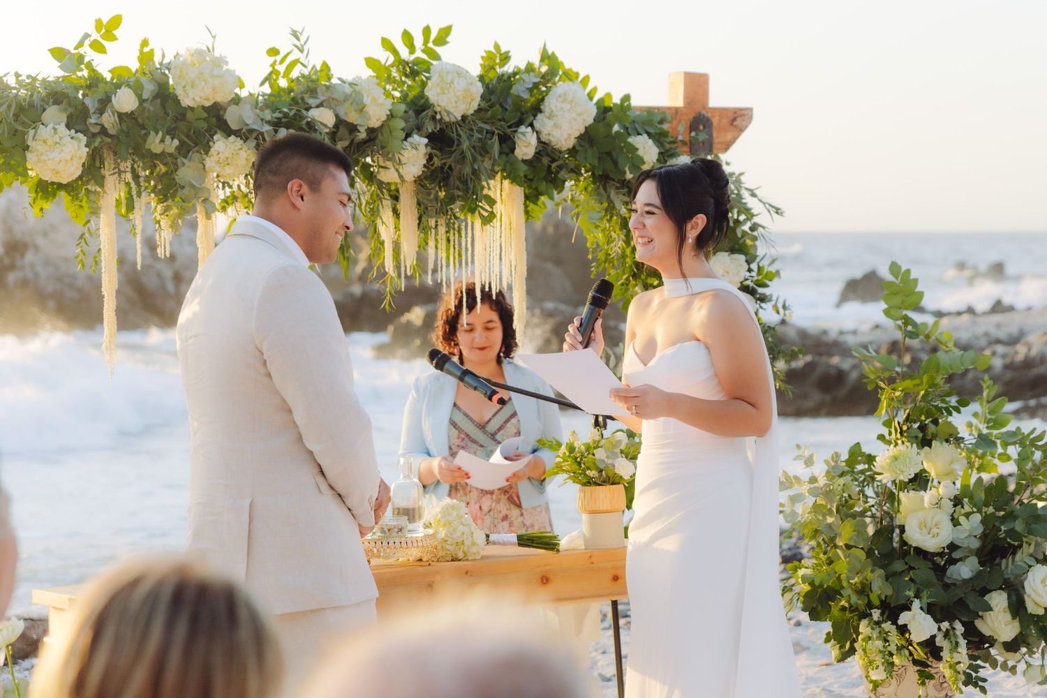 Bride reads vows to groom at romantic beach wedding ceremony with white floral arch and ocean backdrop.