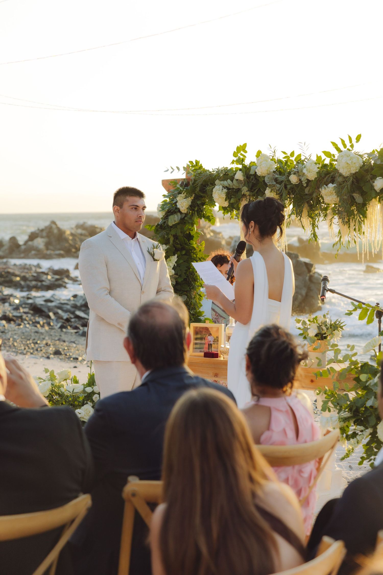 Couple exchanges vows at sunset beach wedding with floral arch, guests seated on sandy shore.