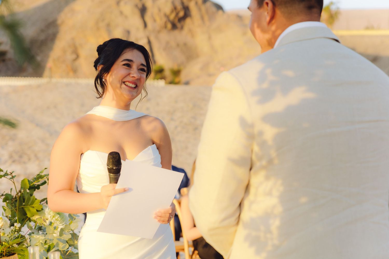 Smiling bride reads vows at outdoor desert wedding ceremony holding paper and microphone.