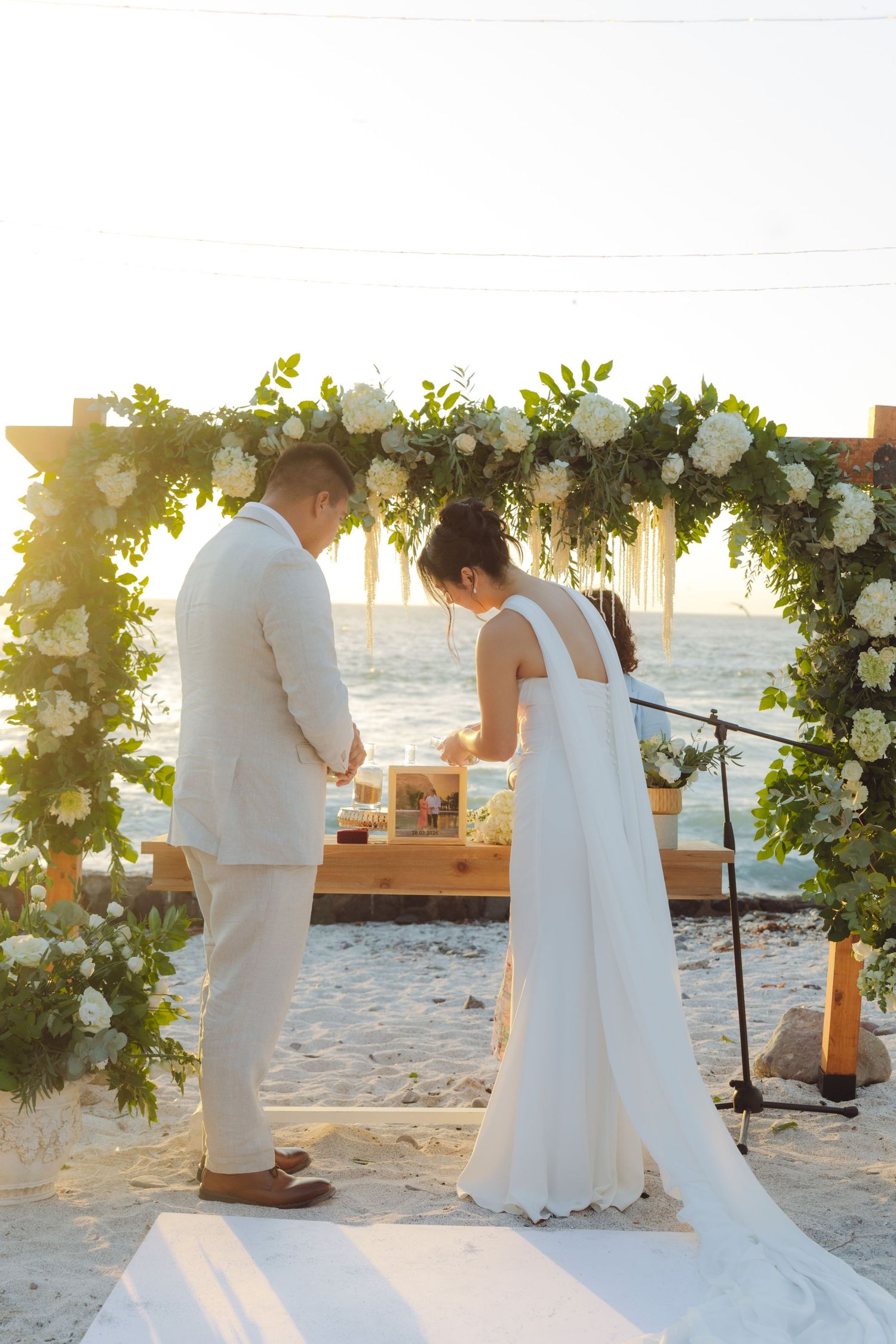 Bride and groom perform sand ceremony at beach wedding under white floral arch at sunset.