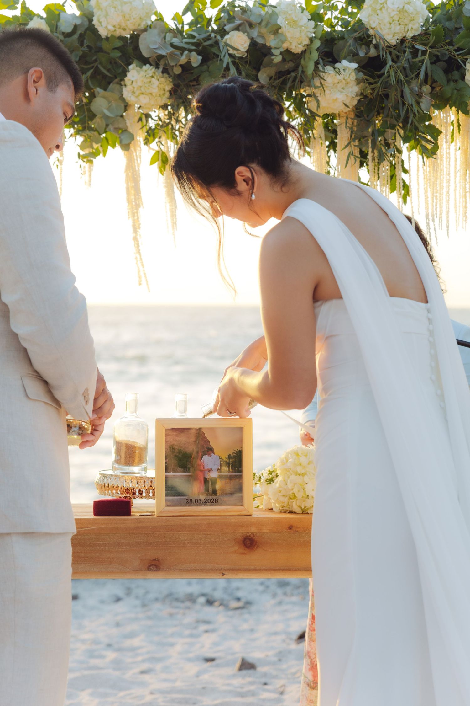Bride pours sand into unity ceremony vessel at beach wedding with floral arch decoration.